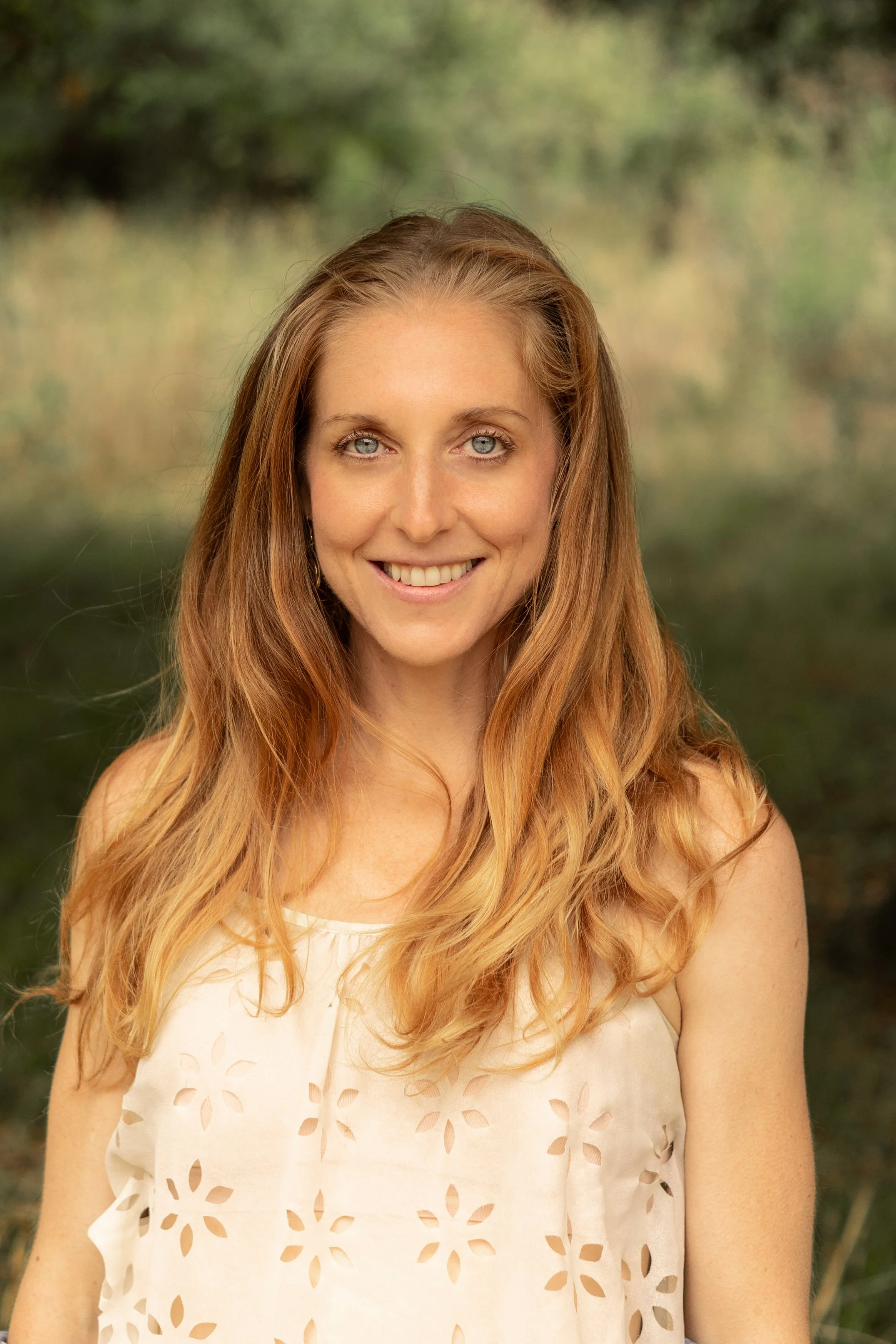 Smiling woman with long light brown hair wearing a white cutout top outdoors