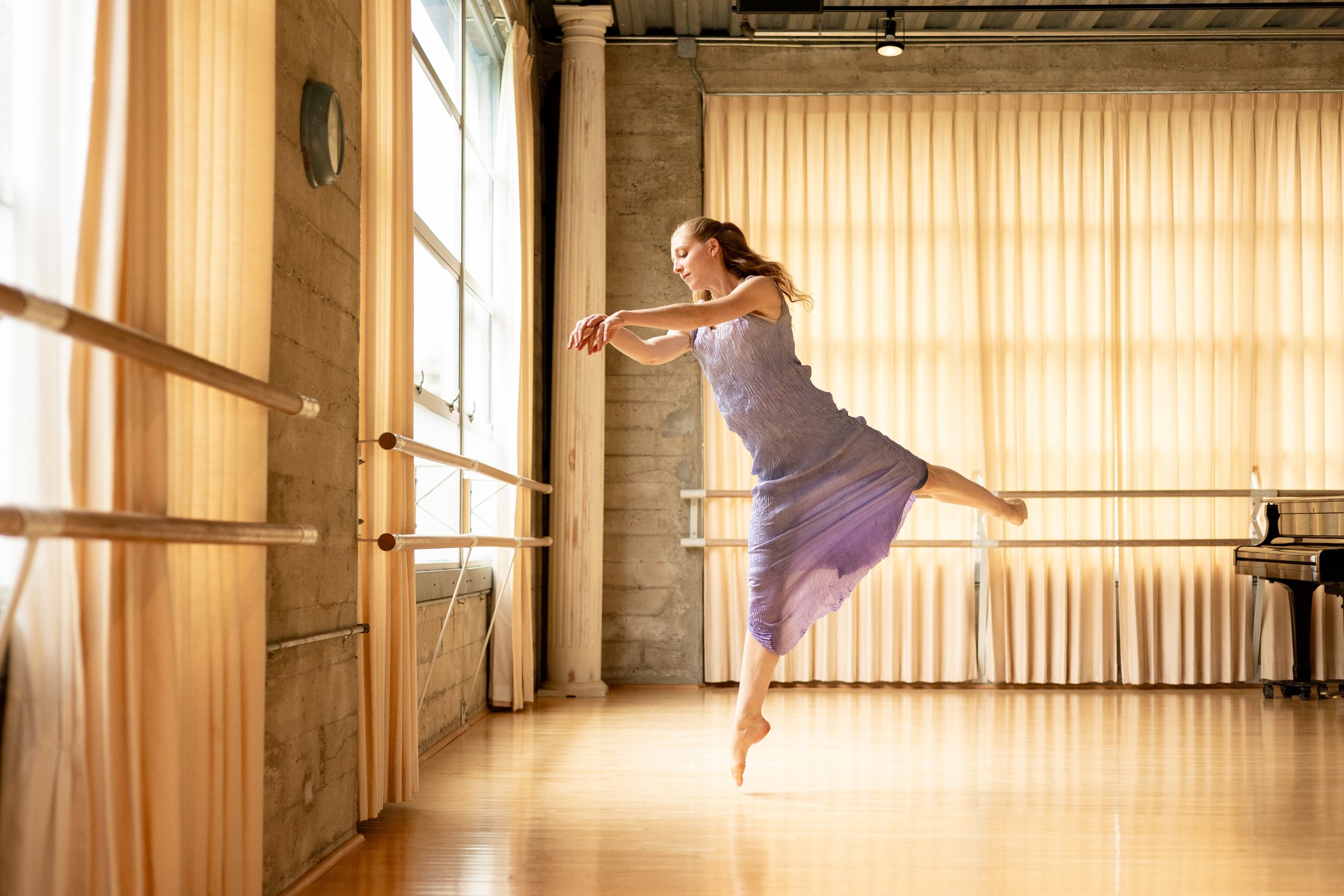 Ballet dancer in a lavender dress performing a leap in a sunlit studio with ballet barres and a grand piano
