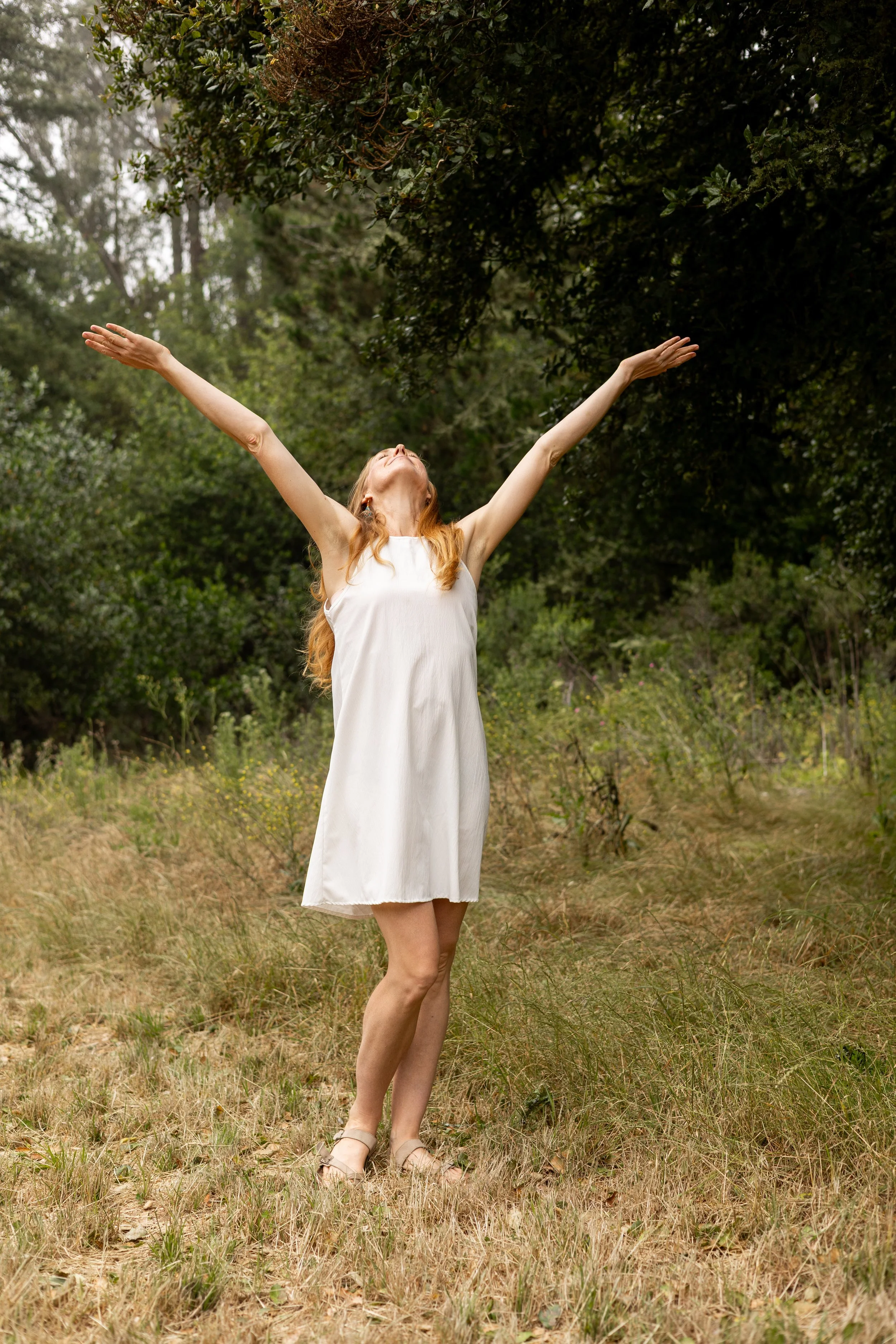 Woman in a white dress standing in a forest clearing with her arms raised.