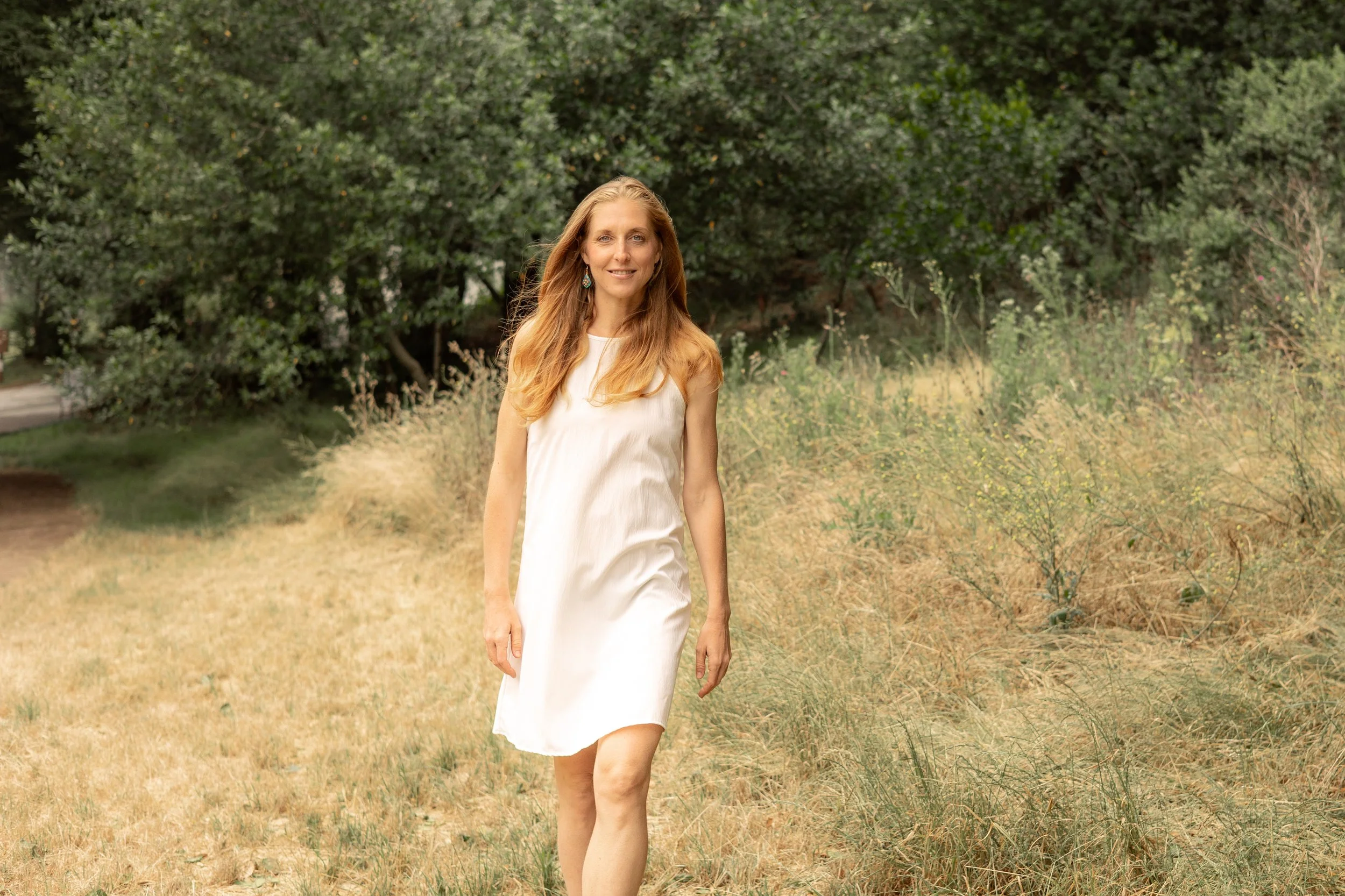 Woman in a white dress walking through a sunlit meadow surrounded by trees