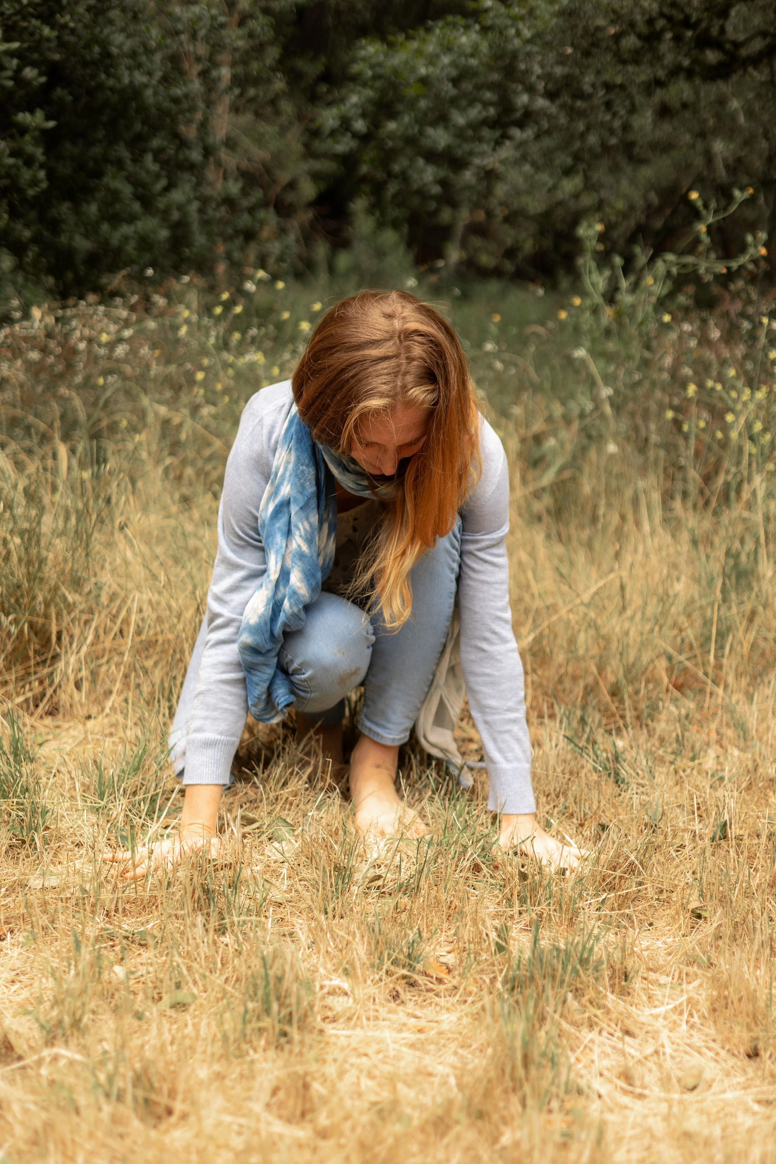 Red-haired woman kneeling in a dry grassy field with a baby in a blue sling
