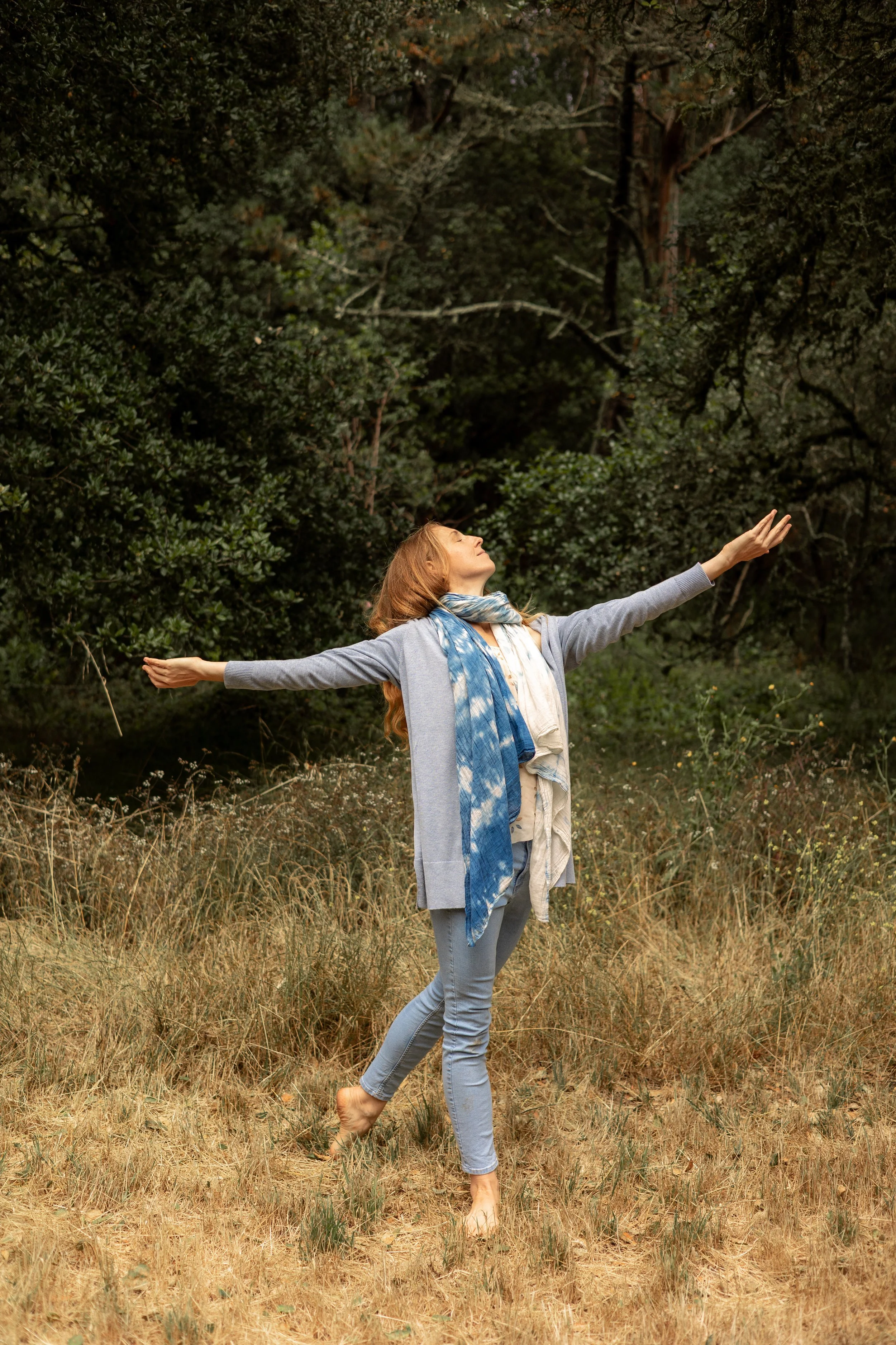 Woman with arms outstretched walking barefoot through a sunlit forest meadow