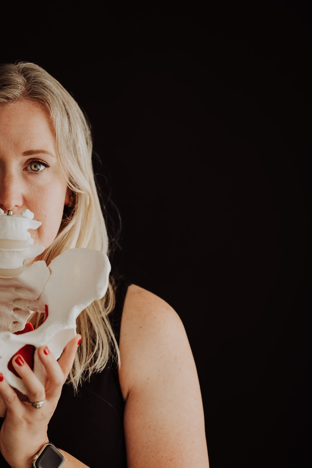 Janelle Kloske, OT and Sex Counselor, holding a pelvis in front of her face, covering her mouth on a black background wearing a black tank top with a serious expression. Only one side of her face is visible