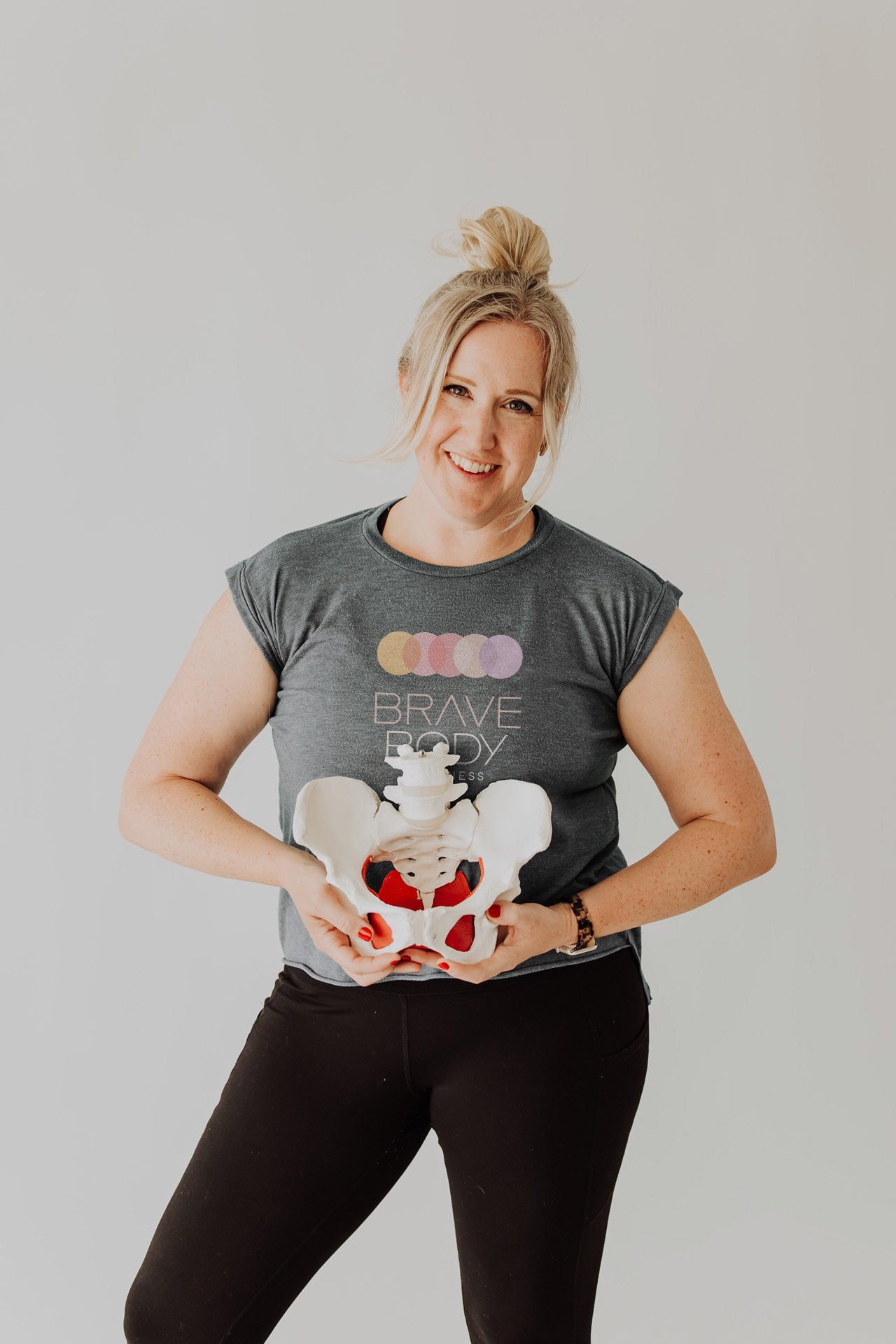 Janelle Kloske, OT and Sex Counselor, smiling and looking at the camera,holding a 3d model of a pelvis, wearing a Brave Body Wellness t-shirt