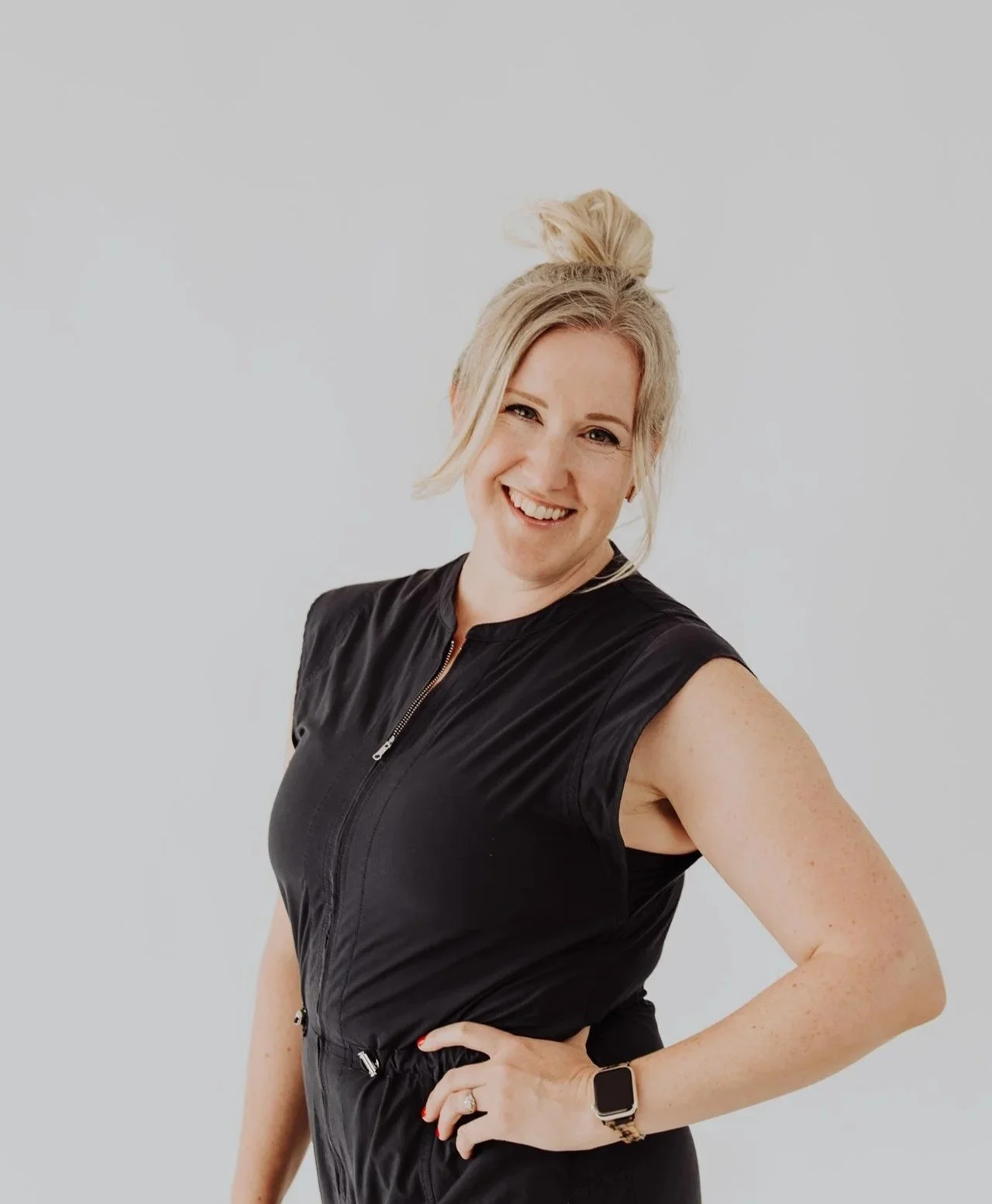 Janelle Kloske, OT and Sex Therapist, wearing a black jumpsuit with one hand on her hip, smiling at the camera, hair up