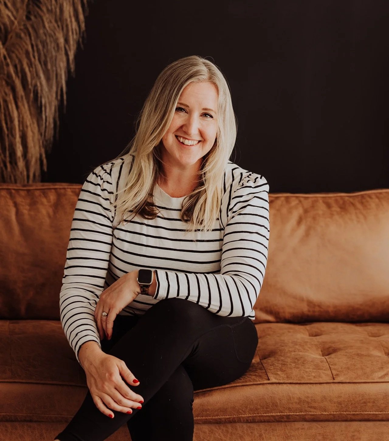 Janelle Kloske, OT and Sex Counselor, sitting on a brown couch, wearing a black and white striped long sleeve shirt, one hand on her lap, legs crossed, looking at the camera and smiling
