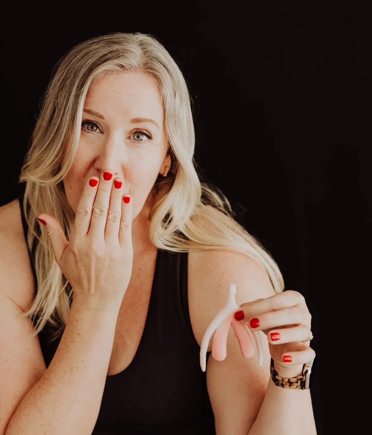Janelle Kloske, OT and Sex Counselor, holding a model of a clitoris in one hand, covering her mouth with the other hand, a mischievous expression on her face. Wearing a black tank top and red nail polish on a black background
