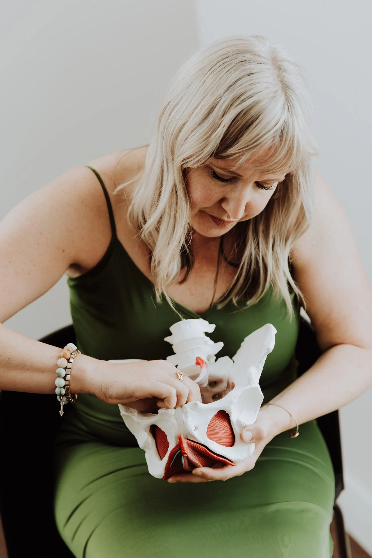 Janelle Kloske, OT and Sex Counselor, holding a 3d model of a pelvis, wearing a green jumpsuit looking down with a serious expression
