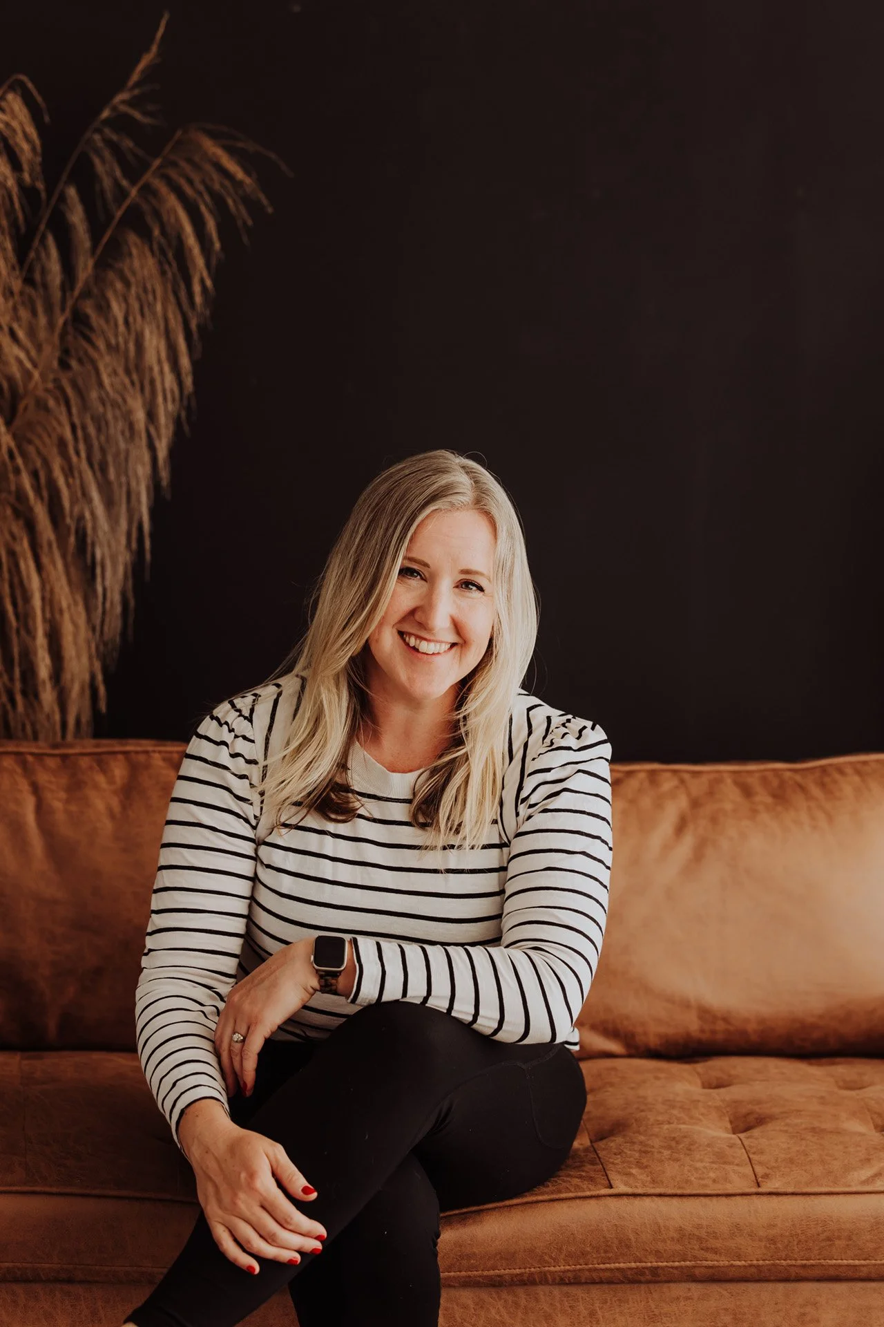 Janelle Kloske, OT and Sex Counselor, sitting on a brown couch, wearing a black and white striped long sleeve shirt, one hand on her lap, legs crossed, looking at the camera and smiling