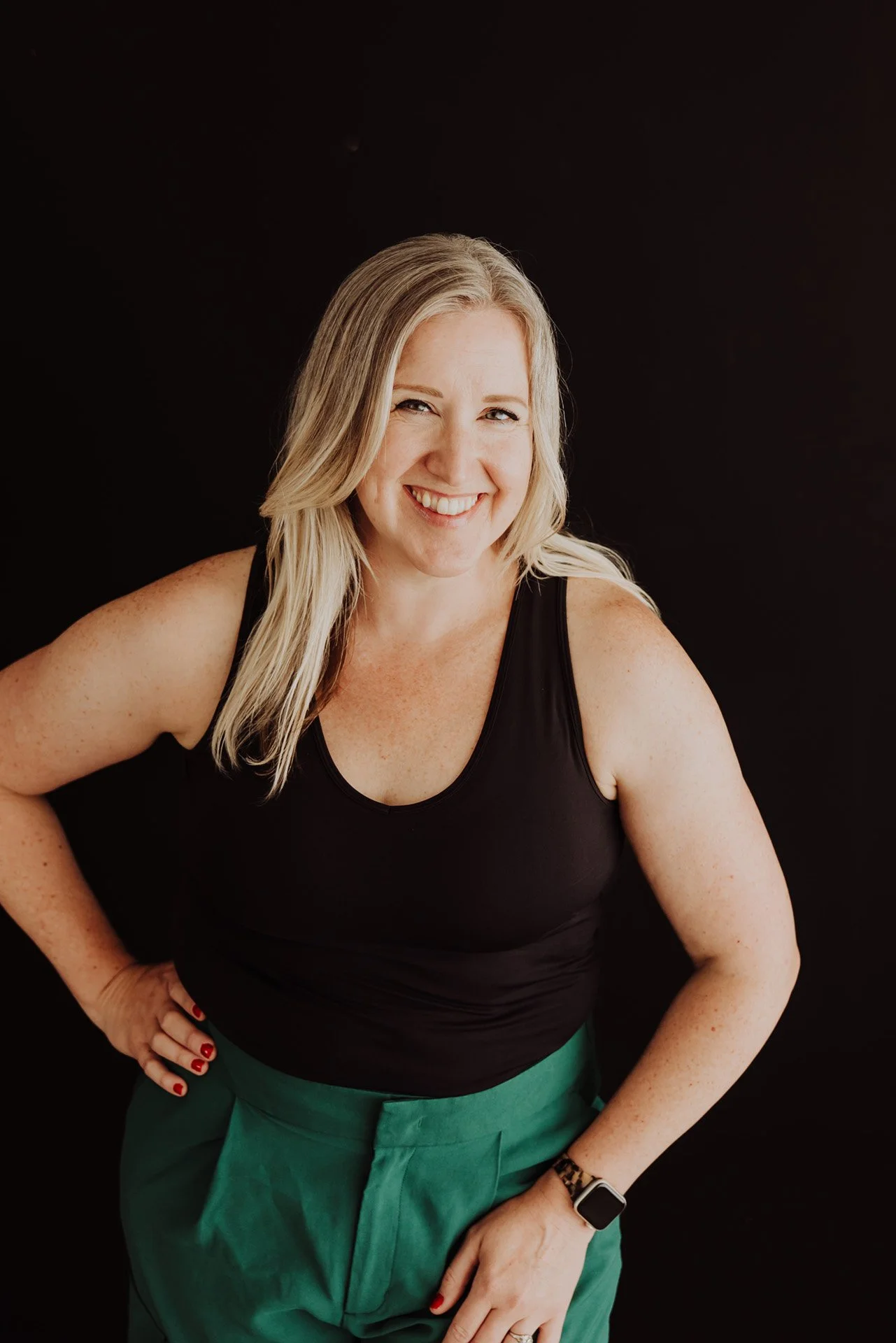 Janelle Kloske, OT and Sex Therapist, dressed in a black tank top and emerald green pants, one hand on her hip, smiling and looking at the camera