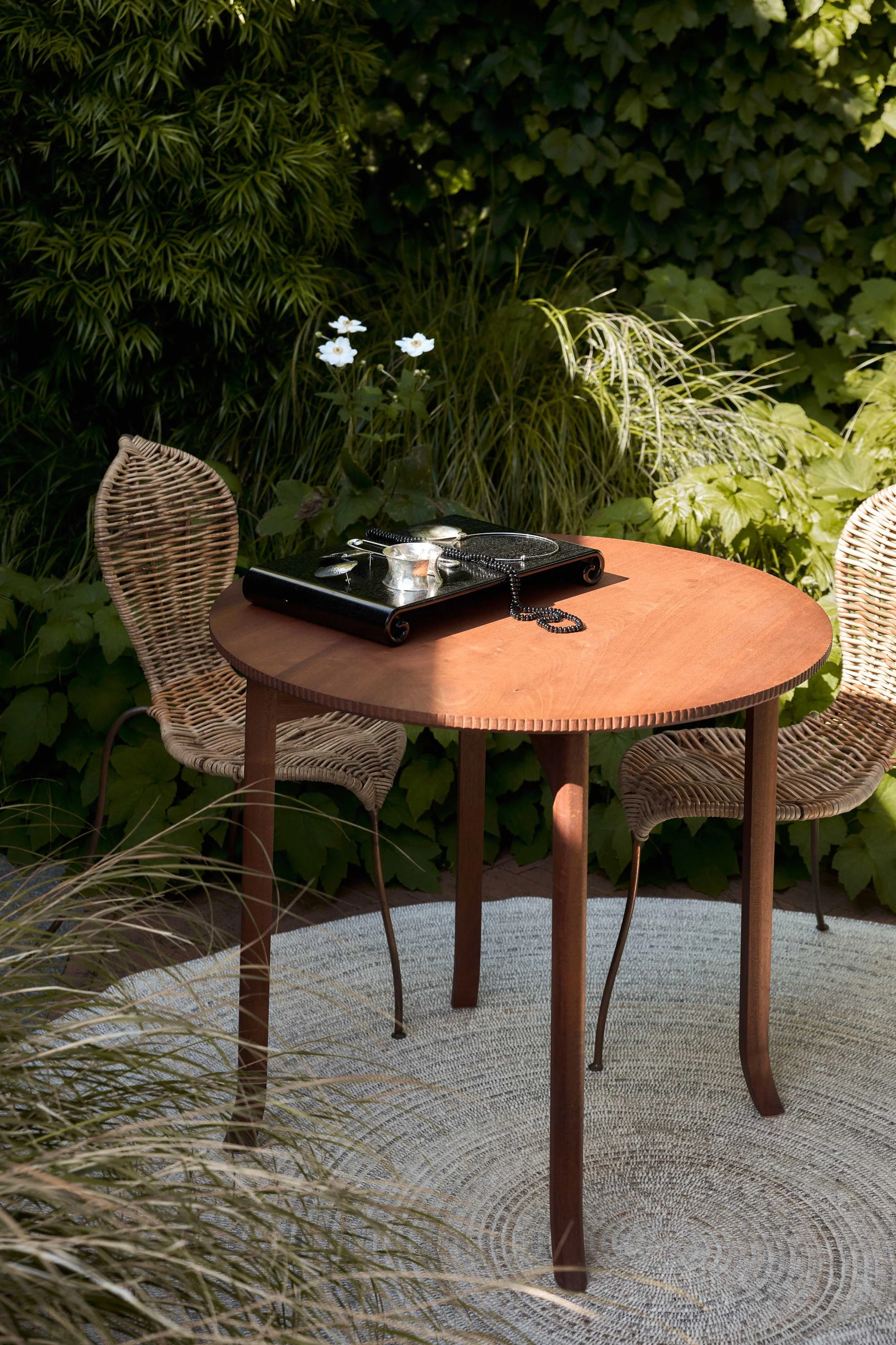 A wooden outdoor table with two wicker chairs surrounded by greenery, with a black tray holding jewelry and a small candle on the table.
