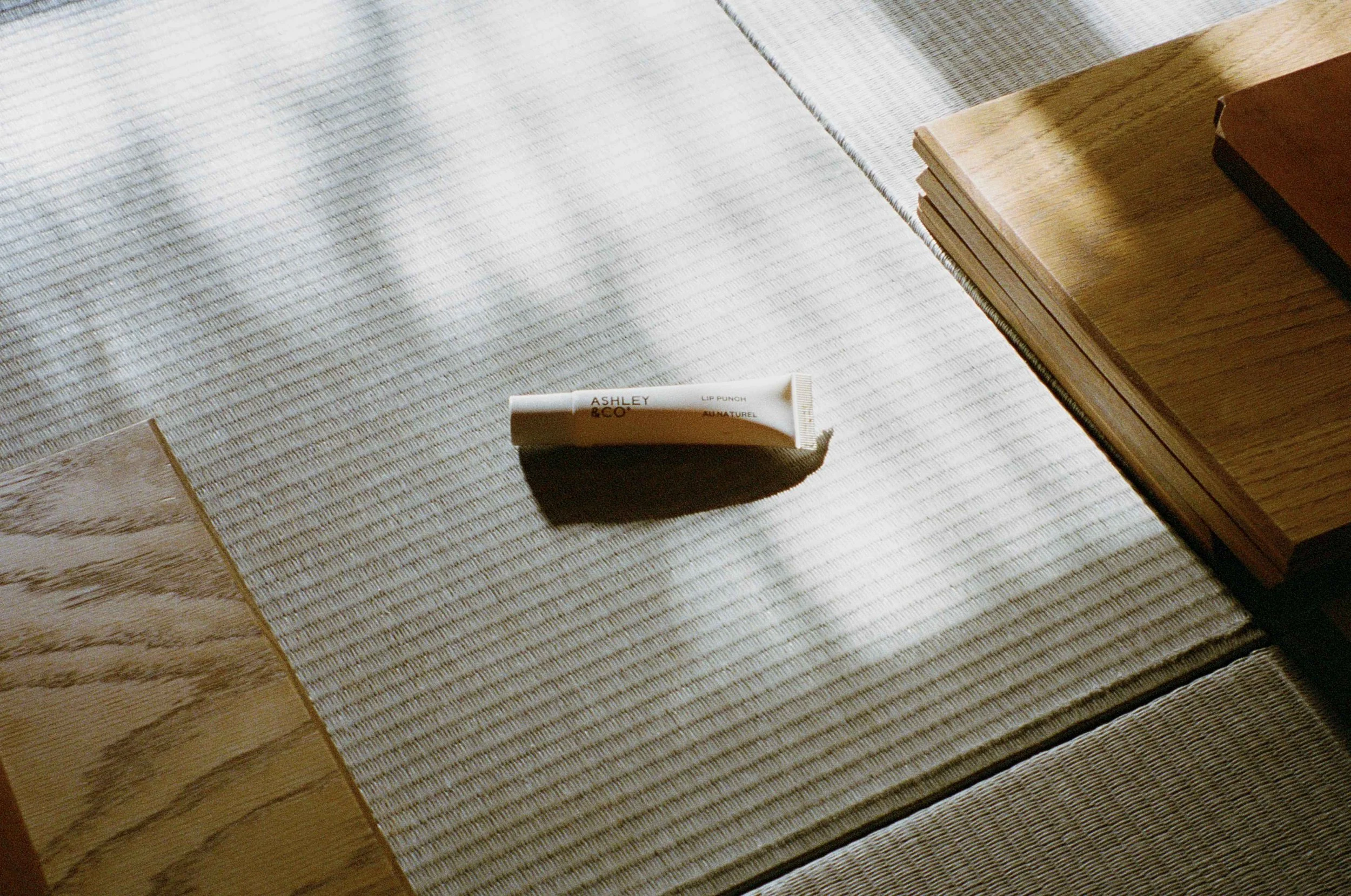 A small white tube labeled 'Ashley & Co Lip Punch All Natural' lies on a textured fabric surface, with shadows cast from a nearby window or curtain. There are wooden surfaces on either side of the fabric.