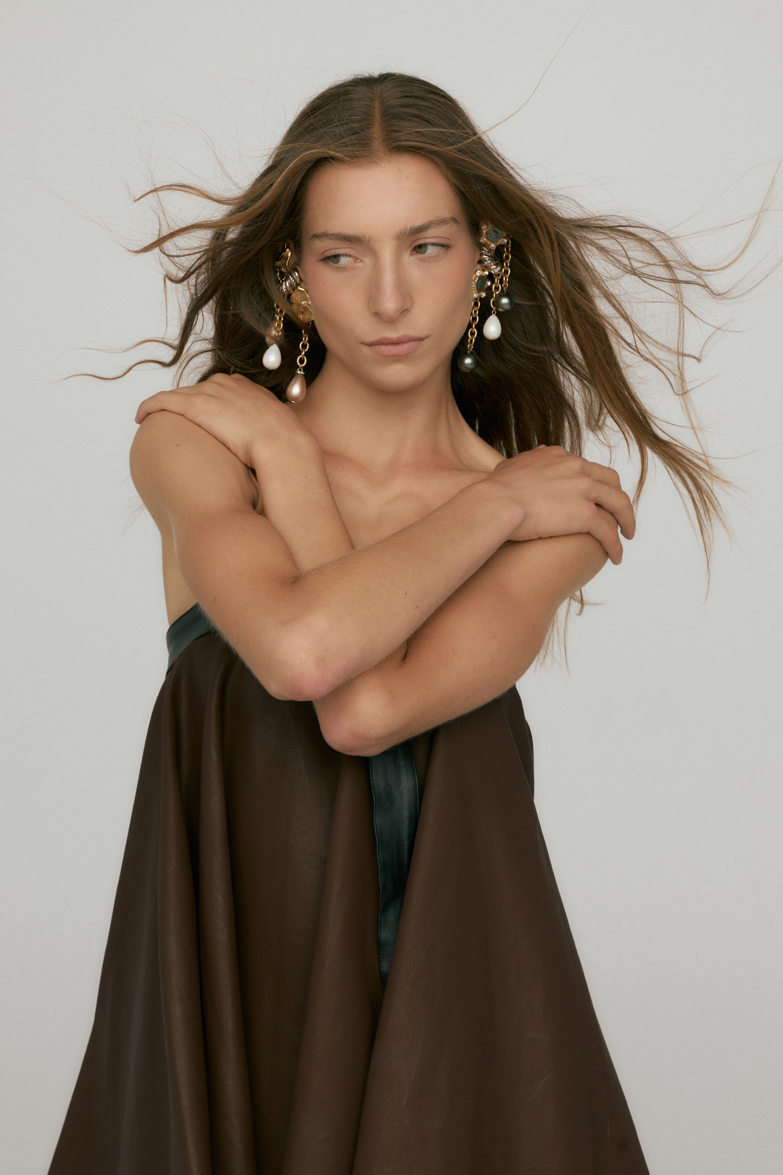 Portrait of a woman with long, flowing hair, wearing multiple earrings, with arms crossed over her shoulders, dressed in a dark brown silk gown, against a plain background.