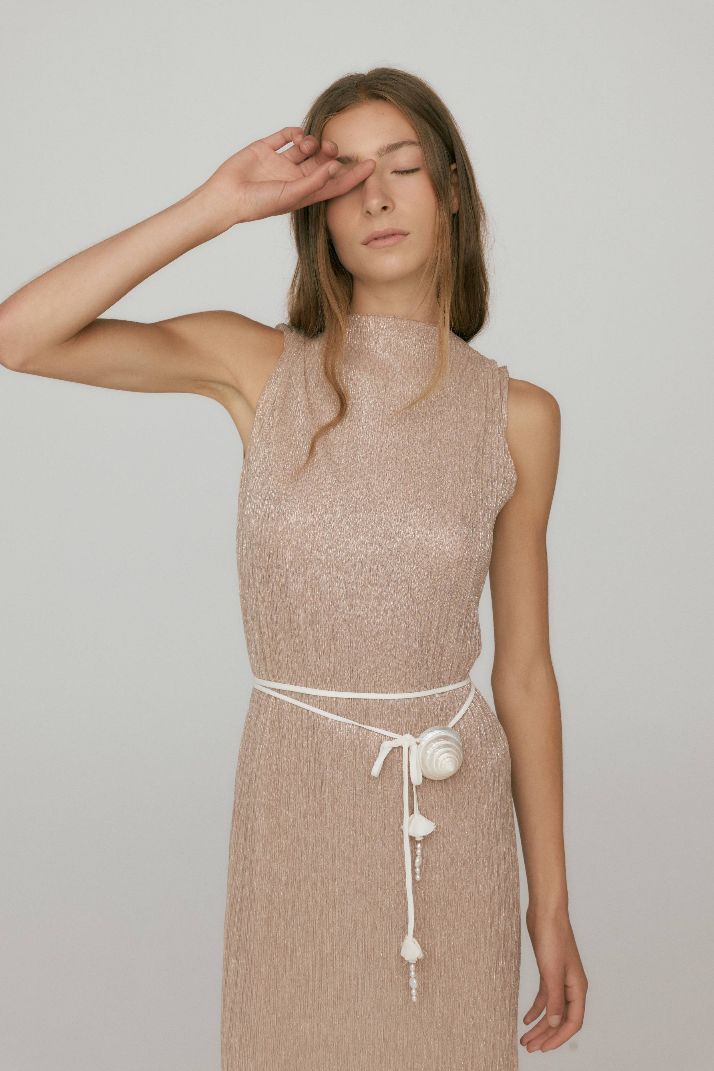 A young woman with long brown hair and closed eyes wearing a sleeveless beige dress, with one hand touching her face, against a plain white background.