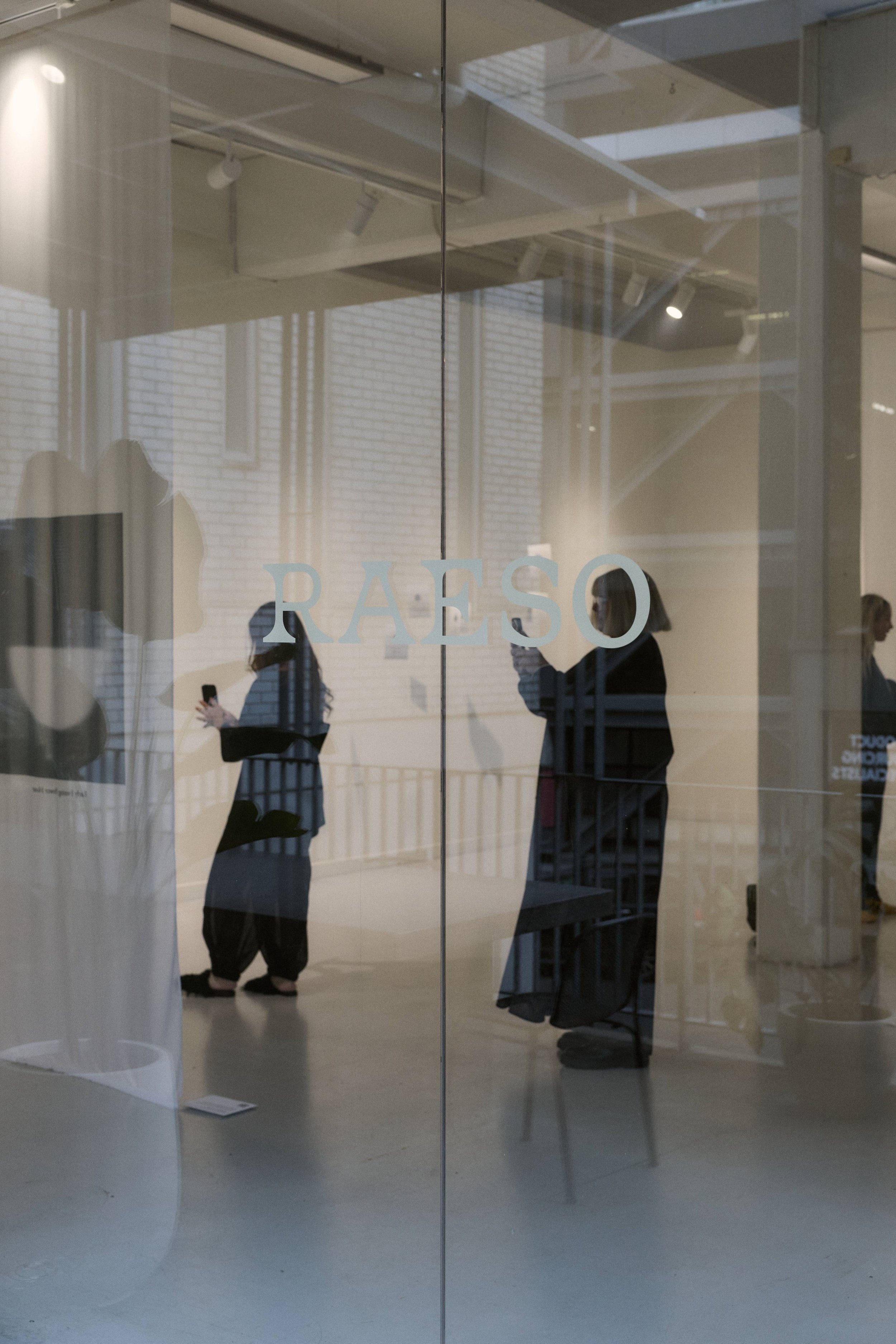 Reflection of people inside an art gallery with the word 'RAESO' on the glass door.