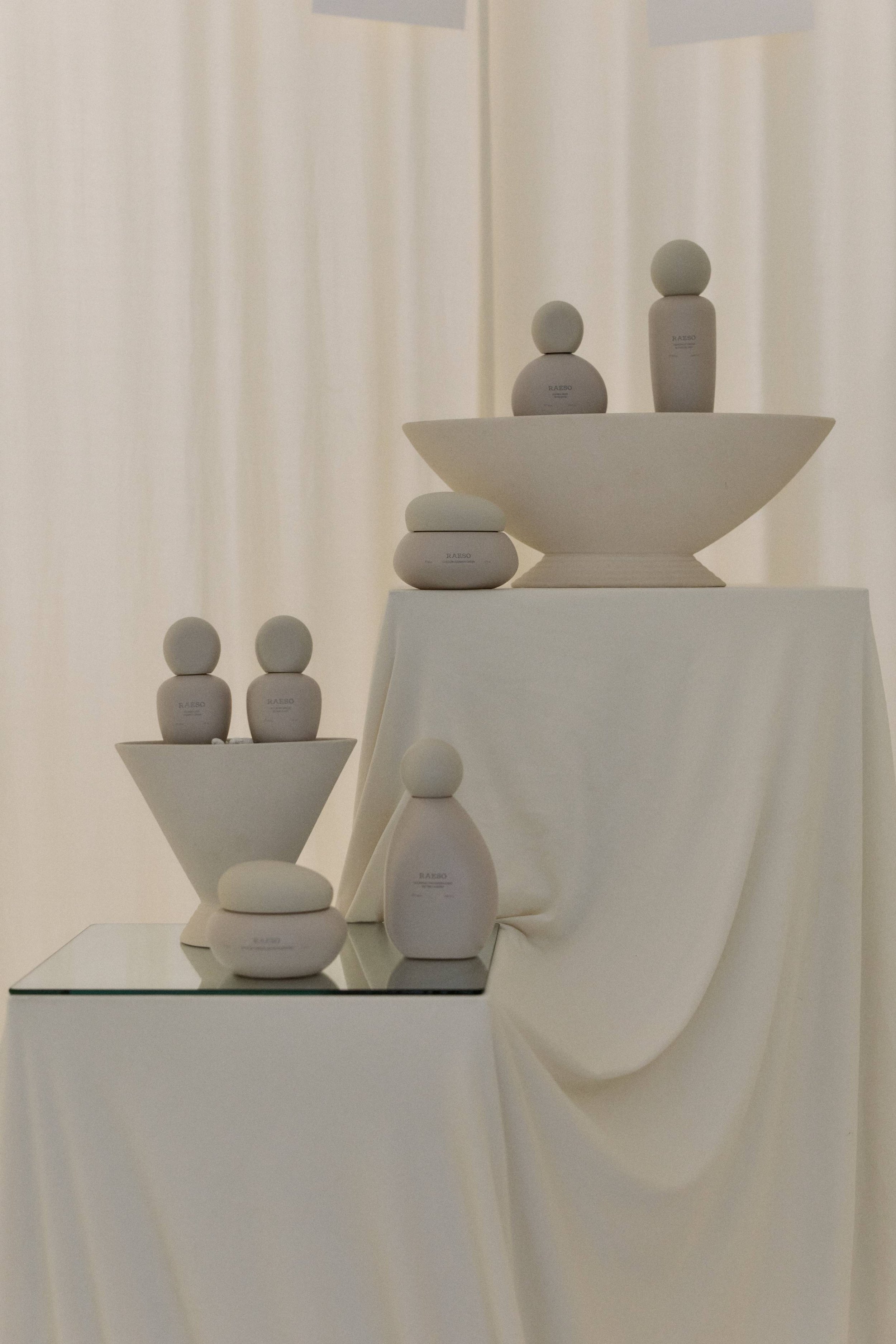 Display of minimalist white ceramic or porcelain figurines arranged on white fabric-covered surfaces with a fabric backdrop.