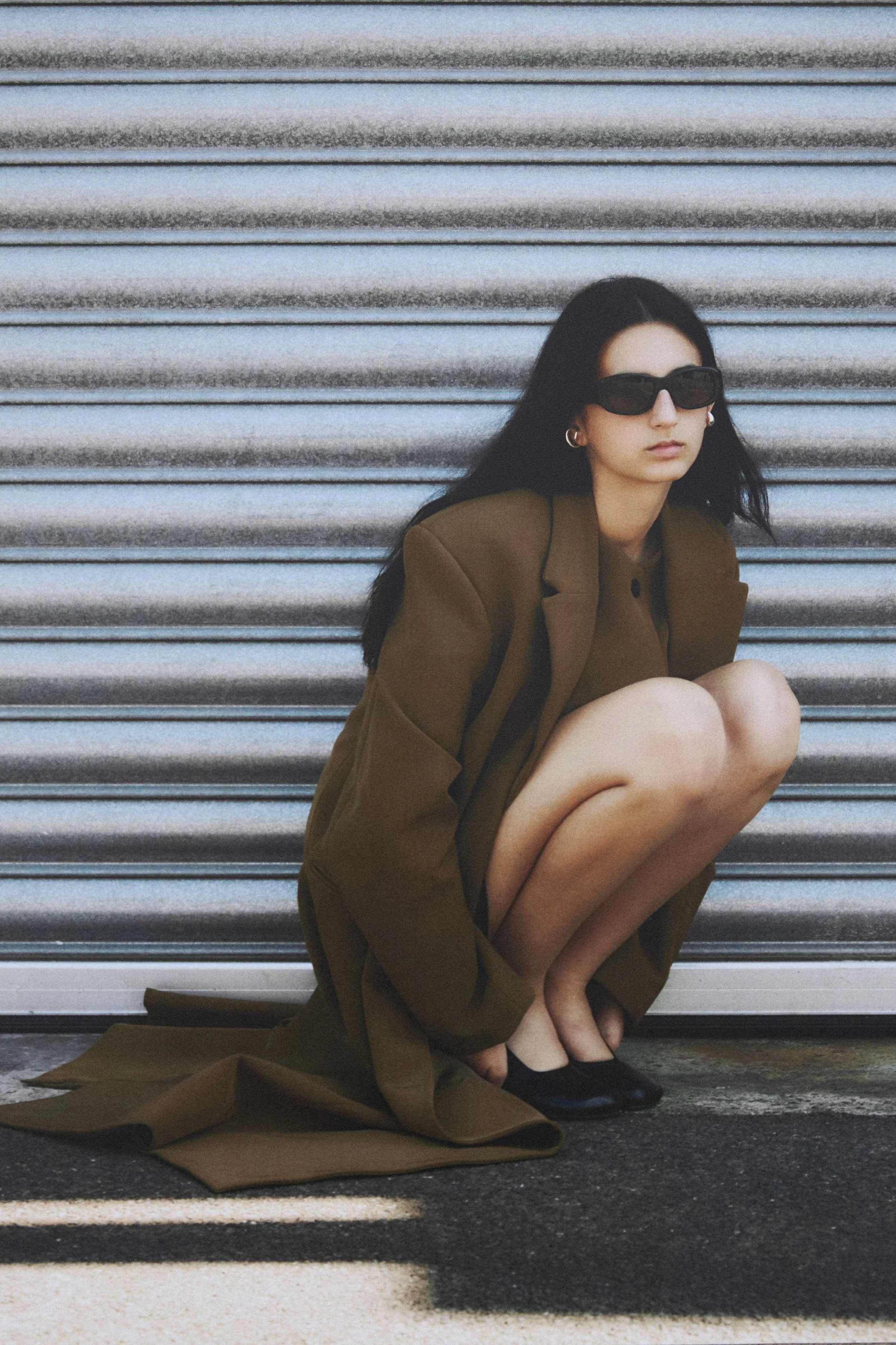A woman crouching in front of a metal rolling shutter, wearing sunglasses, a brown blazer, and black shoes.