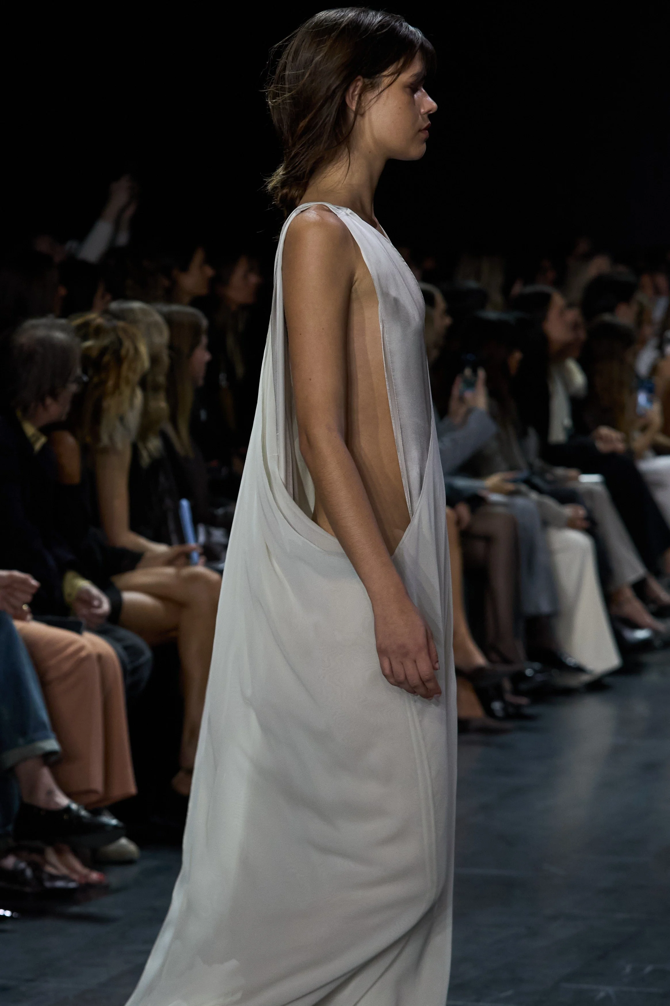 A model walking on a runway during a fashion show, wearing a sleeveless, flowing, gray dress with a sheer panel on the side, with a seated audience in the background.
