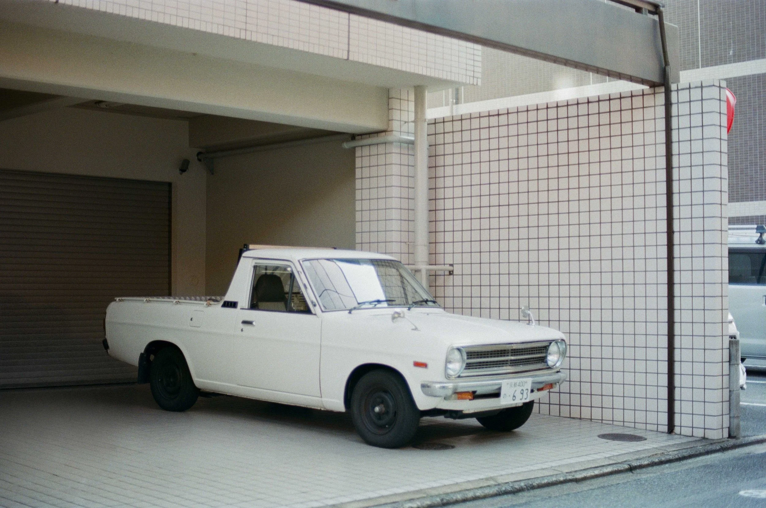 A vintage white pickup truck parked in front of a tiled wall in an urban Japanese setting.