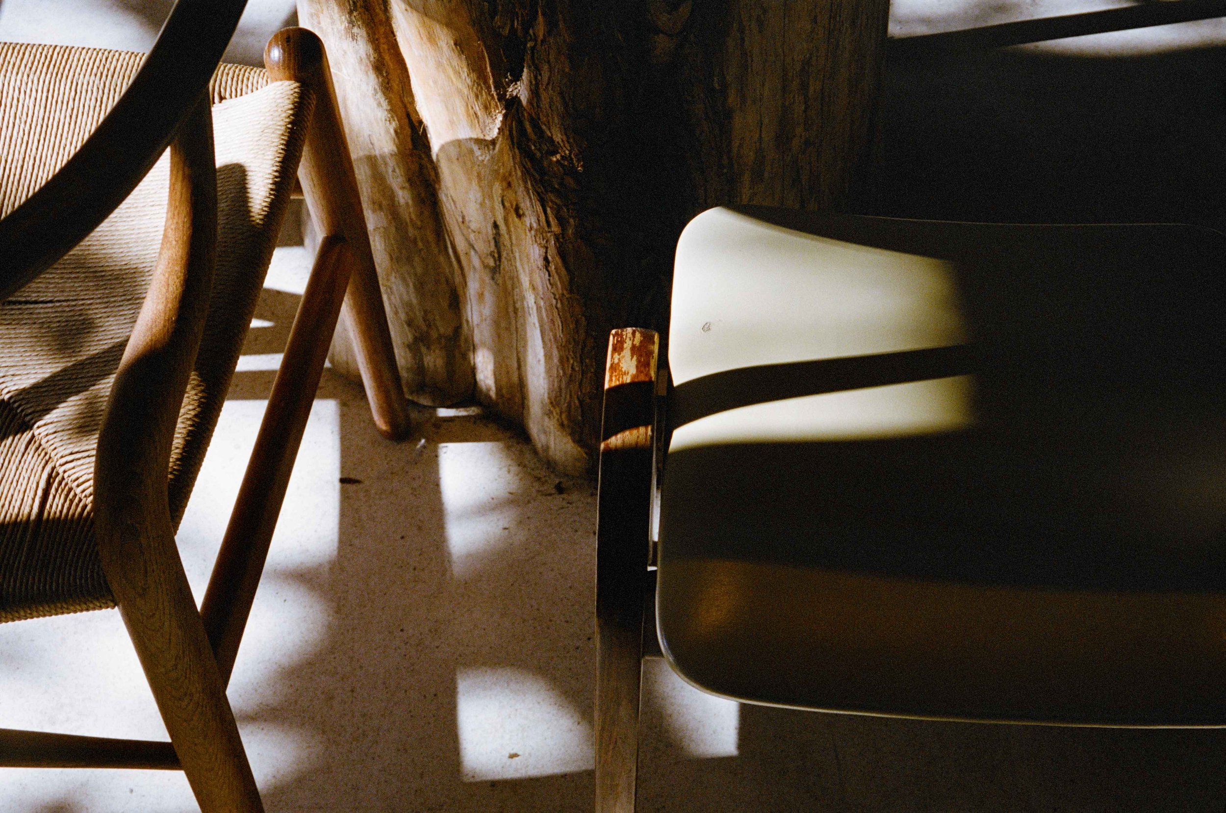 Close-up of the shadow and partial view of three chairs: one with woven rattan and wooden frame, another with a dark metal frame and cream notch seat, and a third with a simple wooden frame, all positioned near a rustic wooden wall and floor.