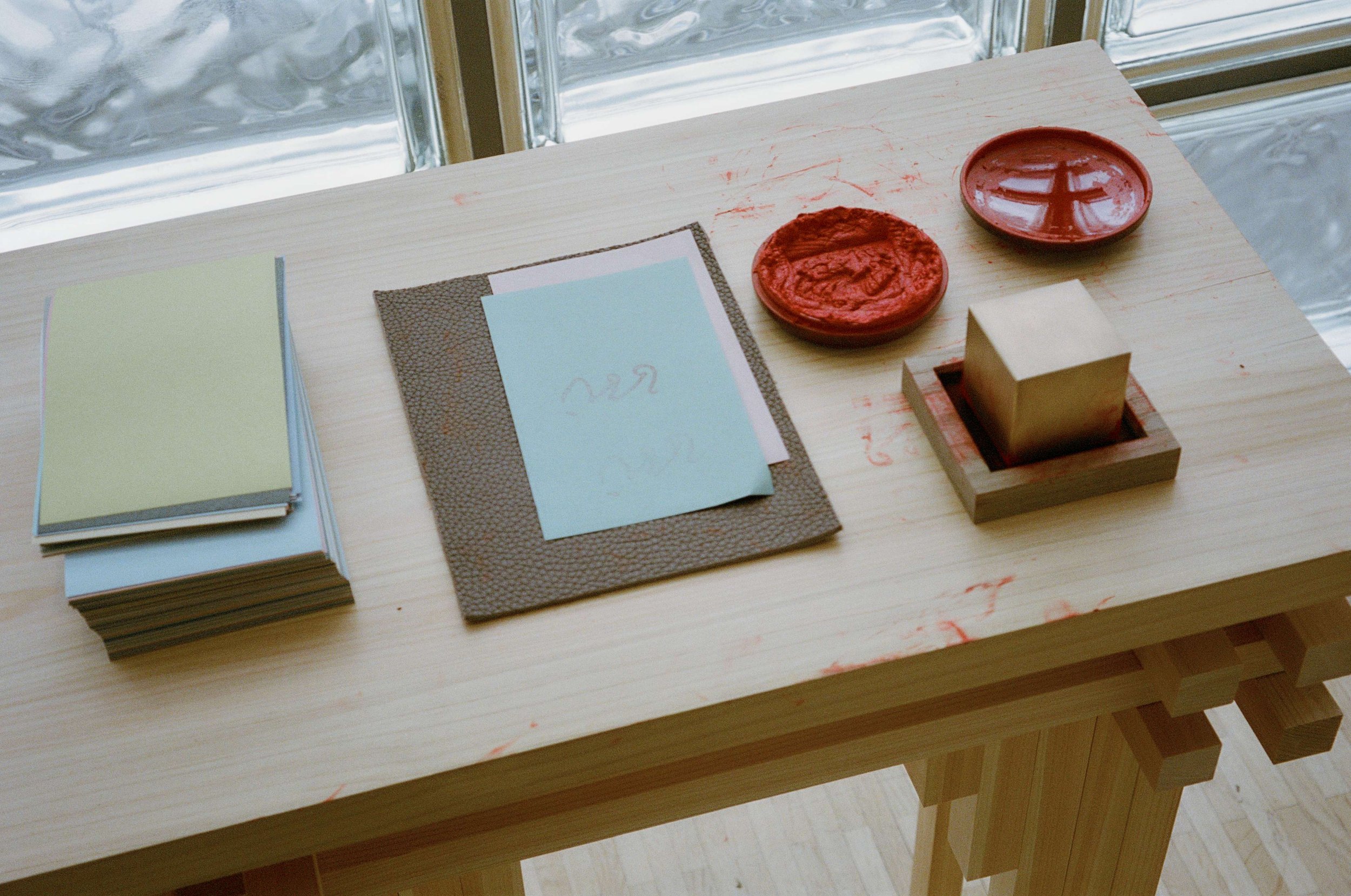 A light-colored wooden table with art supplies including a stack of colored paper, a textured black folder with white and blue papers, a red ink pad, and a gold square stamp in a wooden holder, near a window.