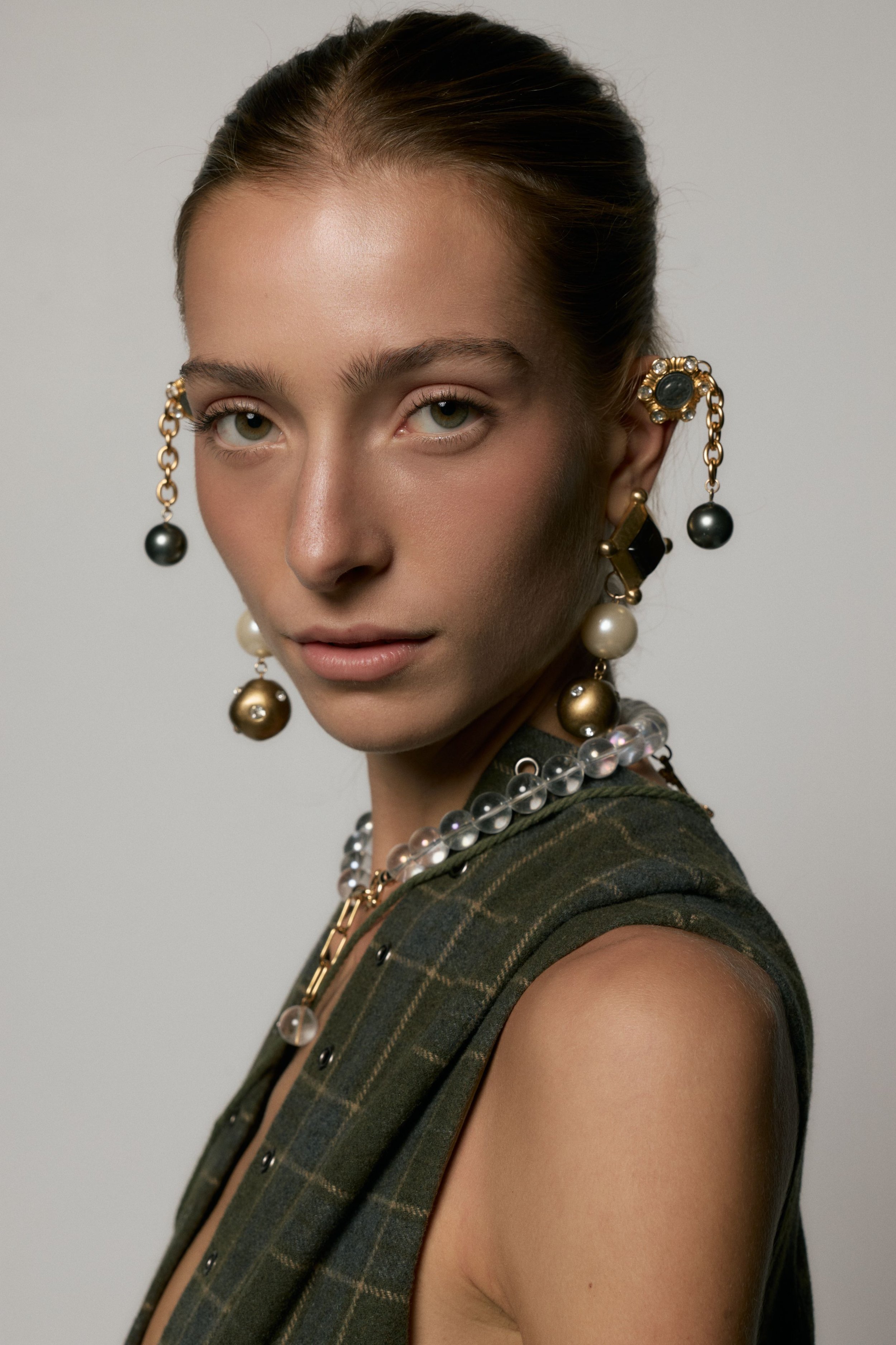 A woman with styled hair and distinctive jewelry including earrings and necklaces, wearing a plaid sleeveless top, looking directly at the camera with a neutral expression.