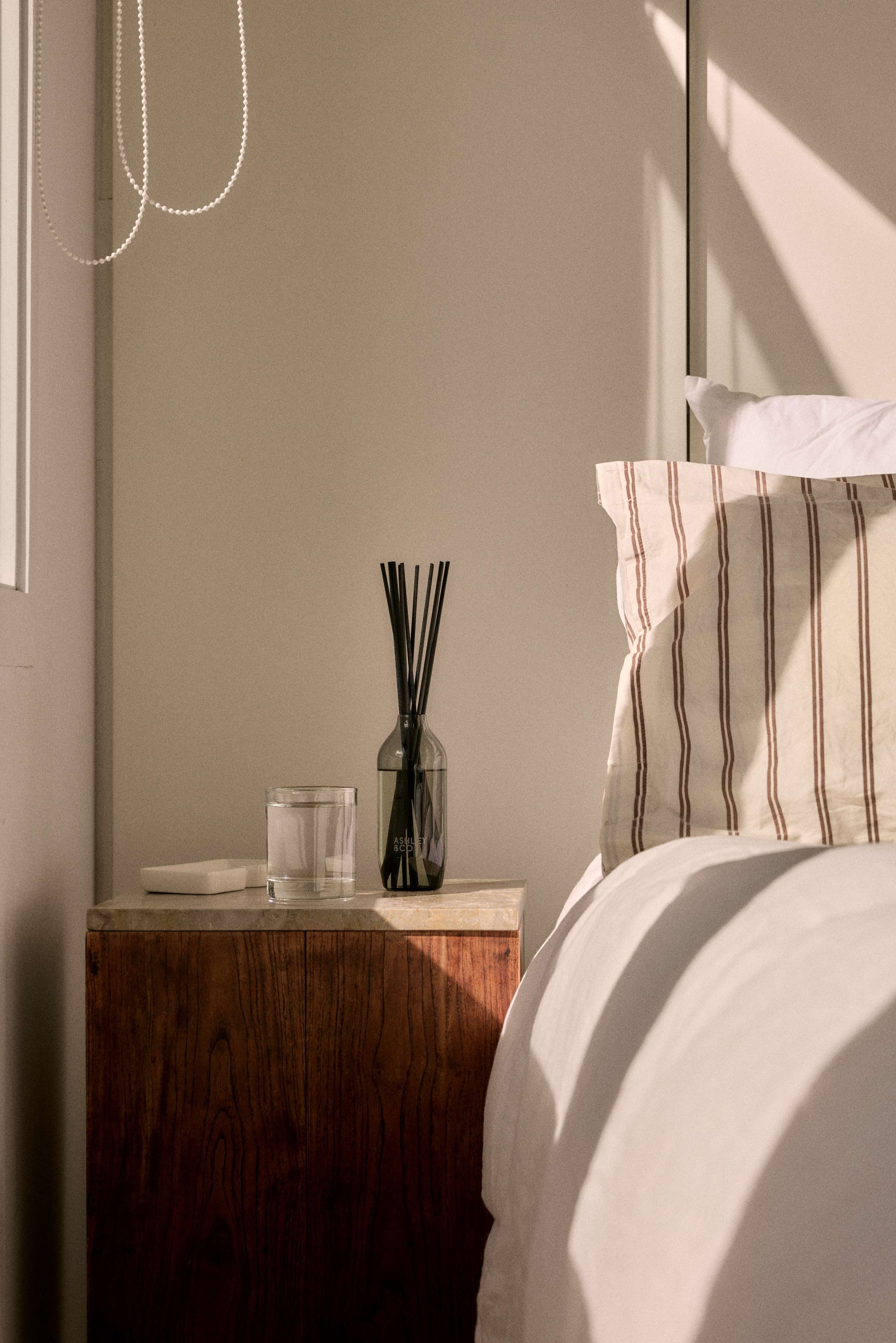A bedroom nightstand with a glass of water, brand name 'Ashley & Co' on the label, a white soap or dish, and a dark reed diffuser container. Part of a bed with white and striped pillows, sunlight casting shadows on the bed and wall.