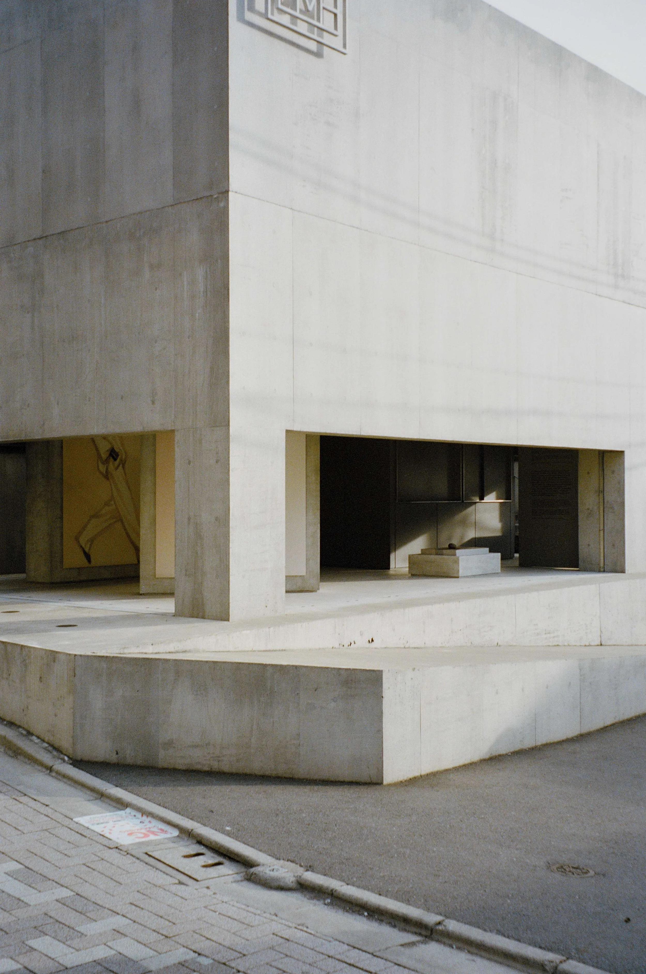 Modern concrete building with an overhanging upper level and shadowed openings on the ground floor, along a brick sidewalk.