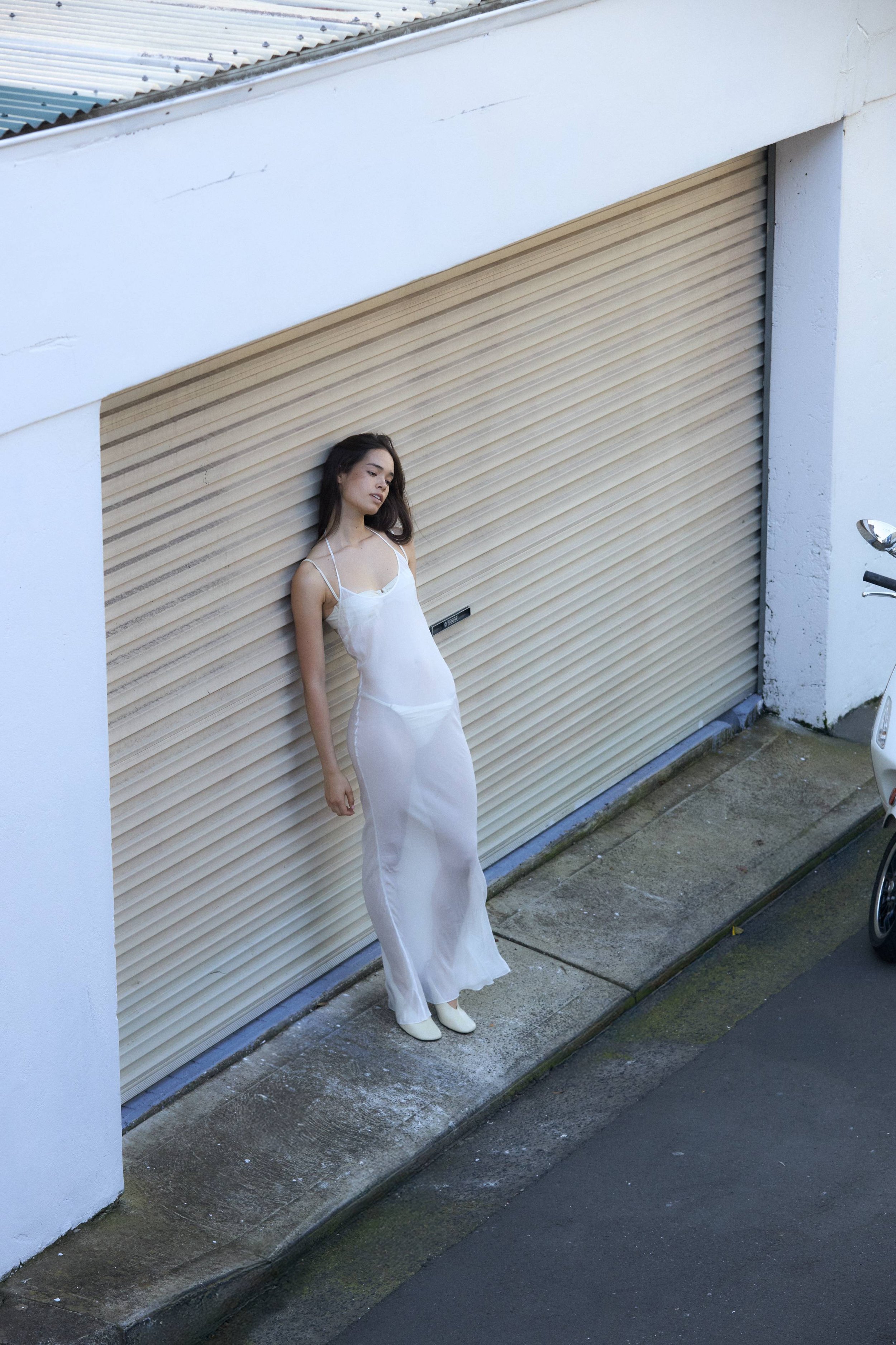 A woman in a white Mise Label satin dress and white shoes standing sideways in front of a beige closed garage door, with a white wall and sidewalk in the background.