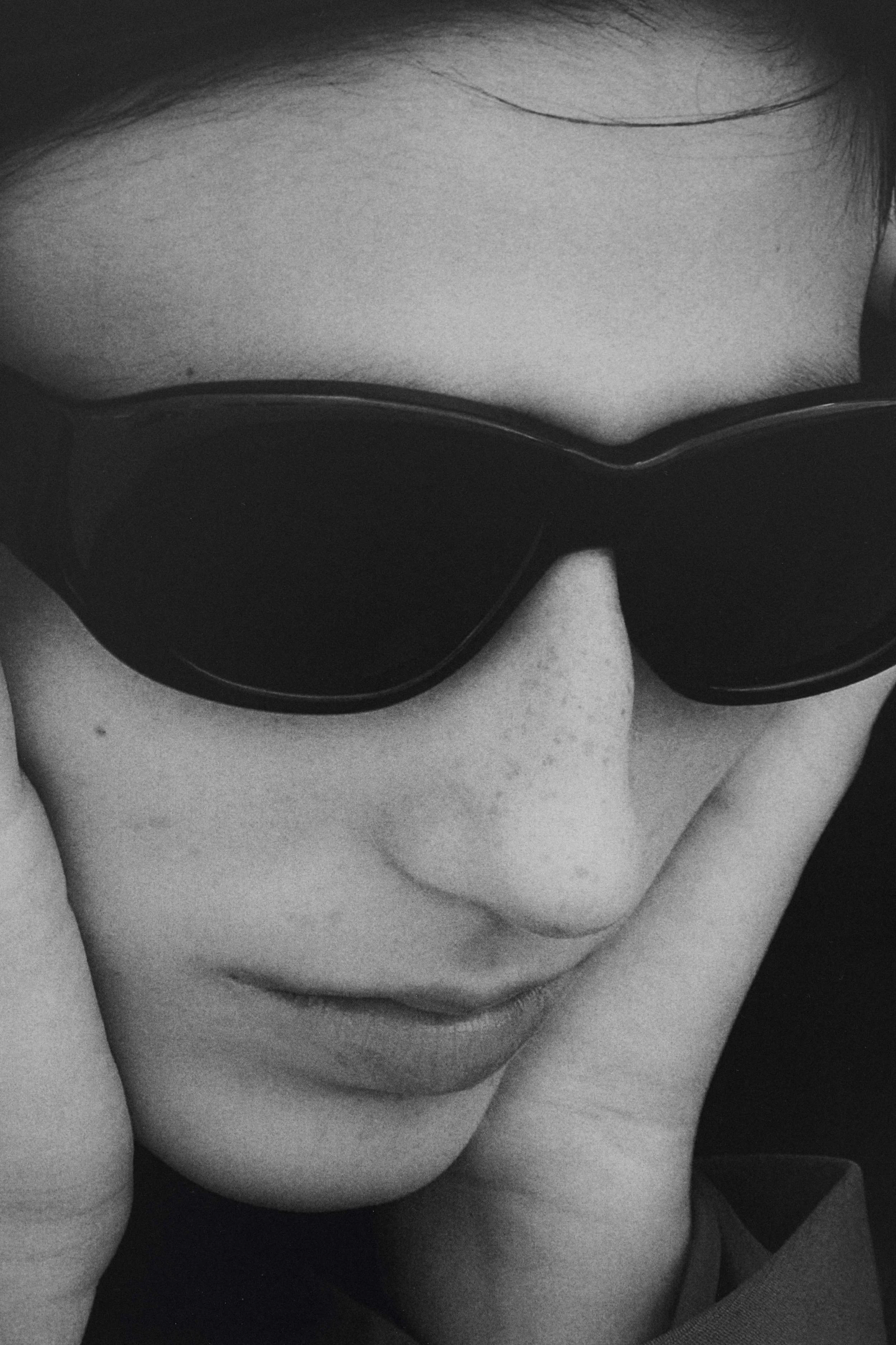 Close-up black-and-white photo of a person's face wearing large sunglasses, viewed from above, with fingers touching the face.