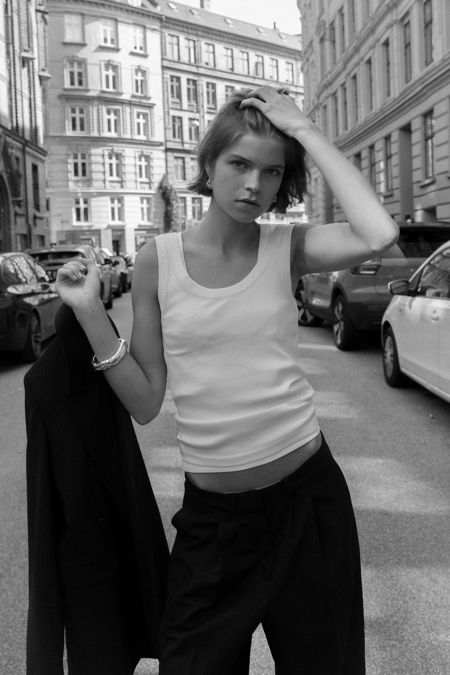 A young woman with short hair in a sleeveless top and dark pants standing on a city street, holding a jacket over her shoulder, with buildings and parked cars in the background.