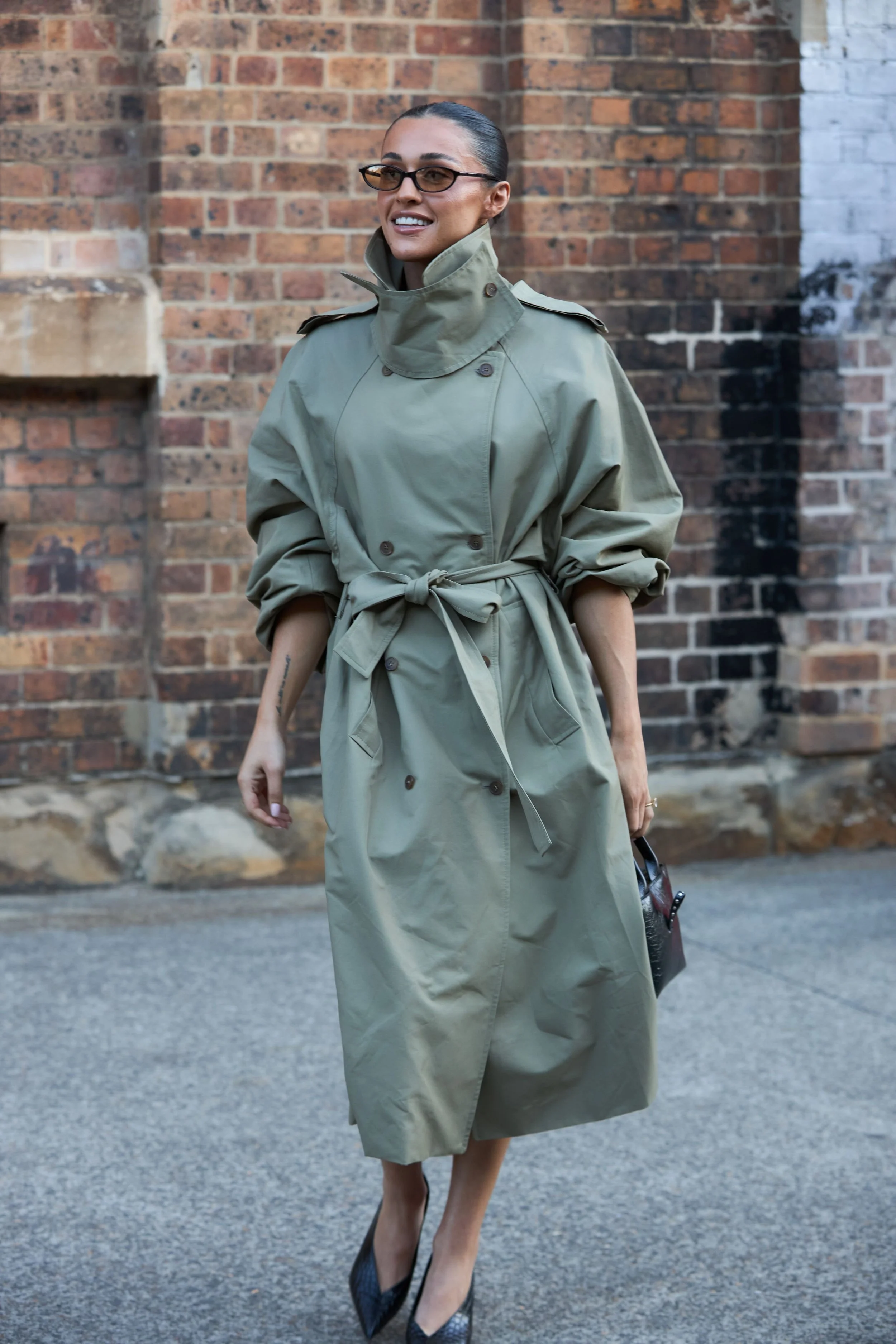Ariana Tapsell wearing a stylish khaki trench coat with rolled-up sleeves and black high heels, standing outdoors against a brick wall with stone accents.