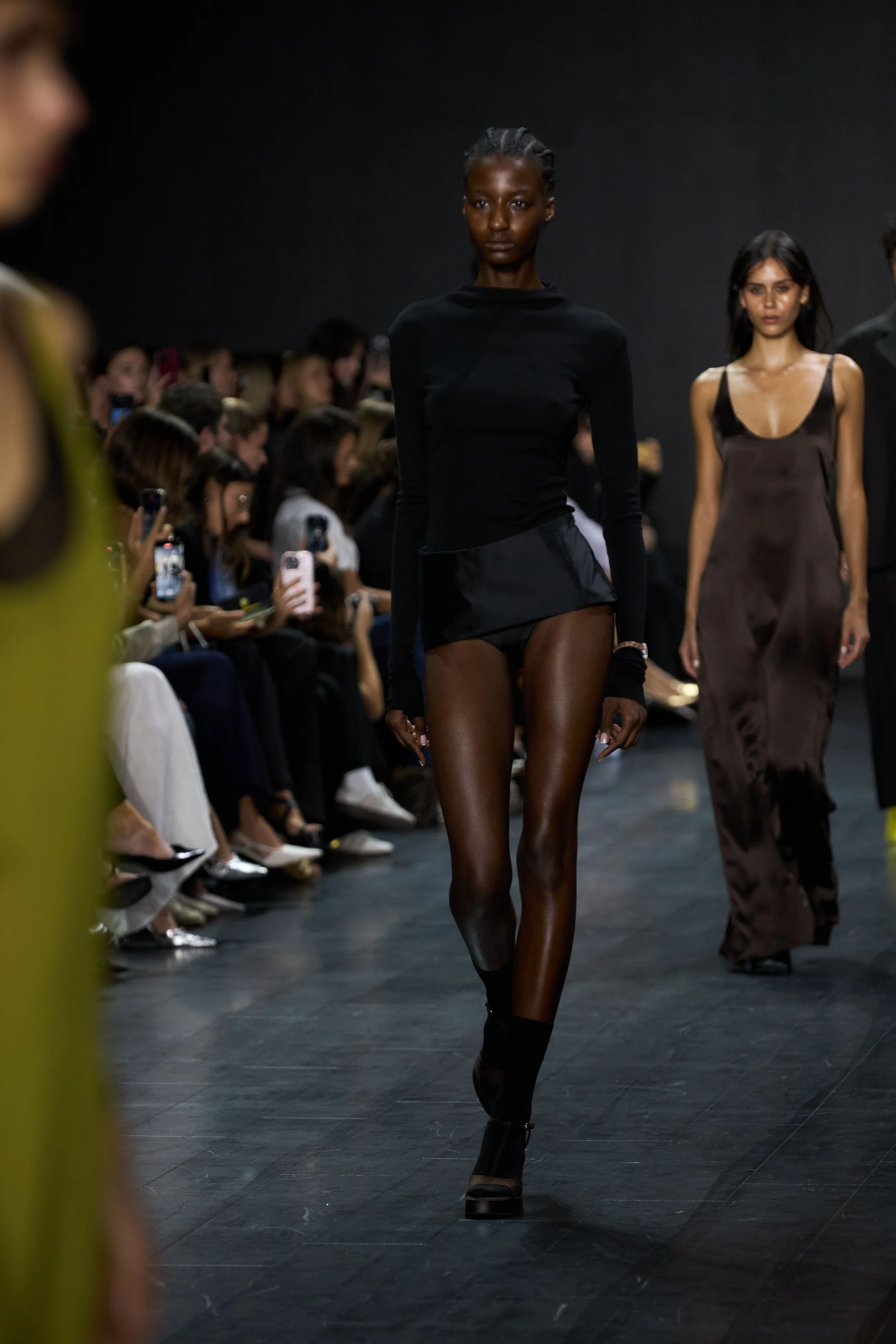 Model walking on runway at a fashion show, wearing black long-sleeve top and black shorts, with audience taking photos for Fashion Quarterly.