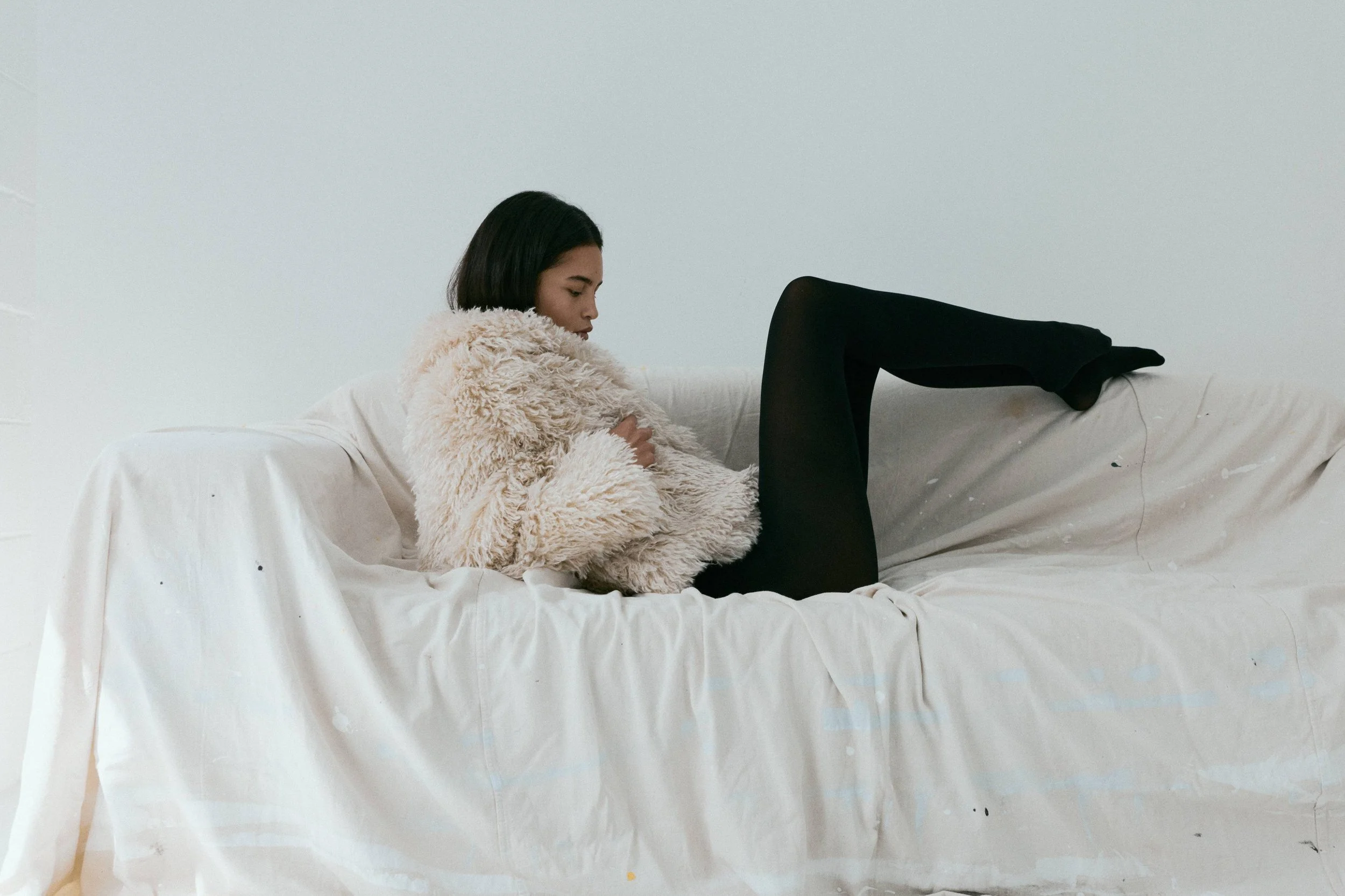 Young woman with short black hair wearing a beige fuzzy coat and black tights relaxing on a white sofa against a plain light-colored wall.