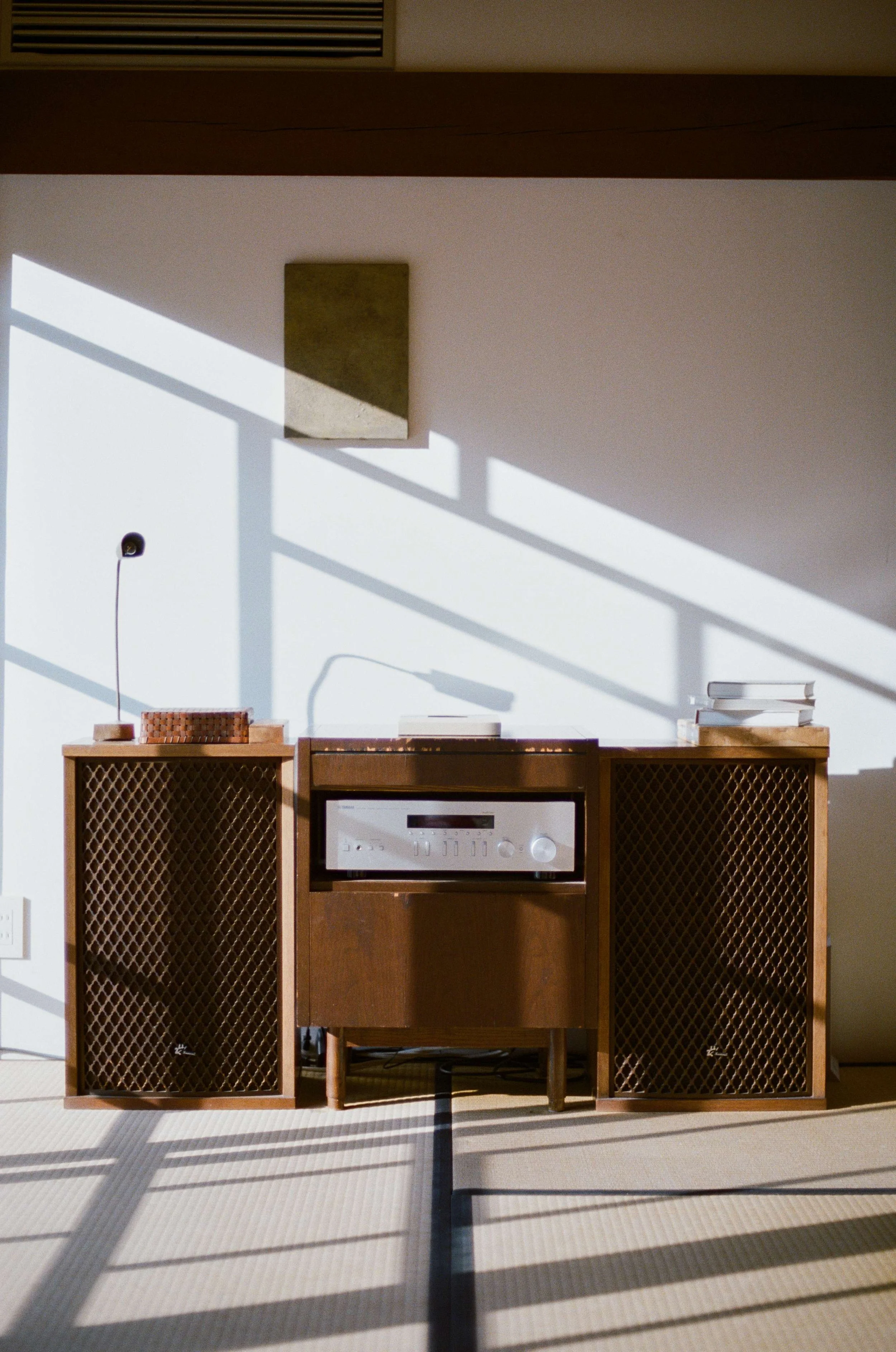 A vintage wooden Japanese stereo cabinet with a stereo system in the middle, positioned against a white wall with sunlight and shadows cast from large window panes, and a small framed artwork above it.