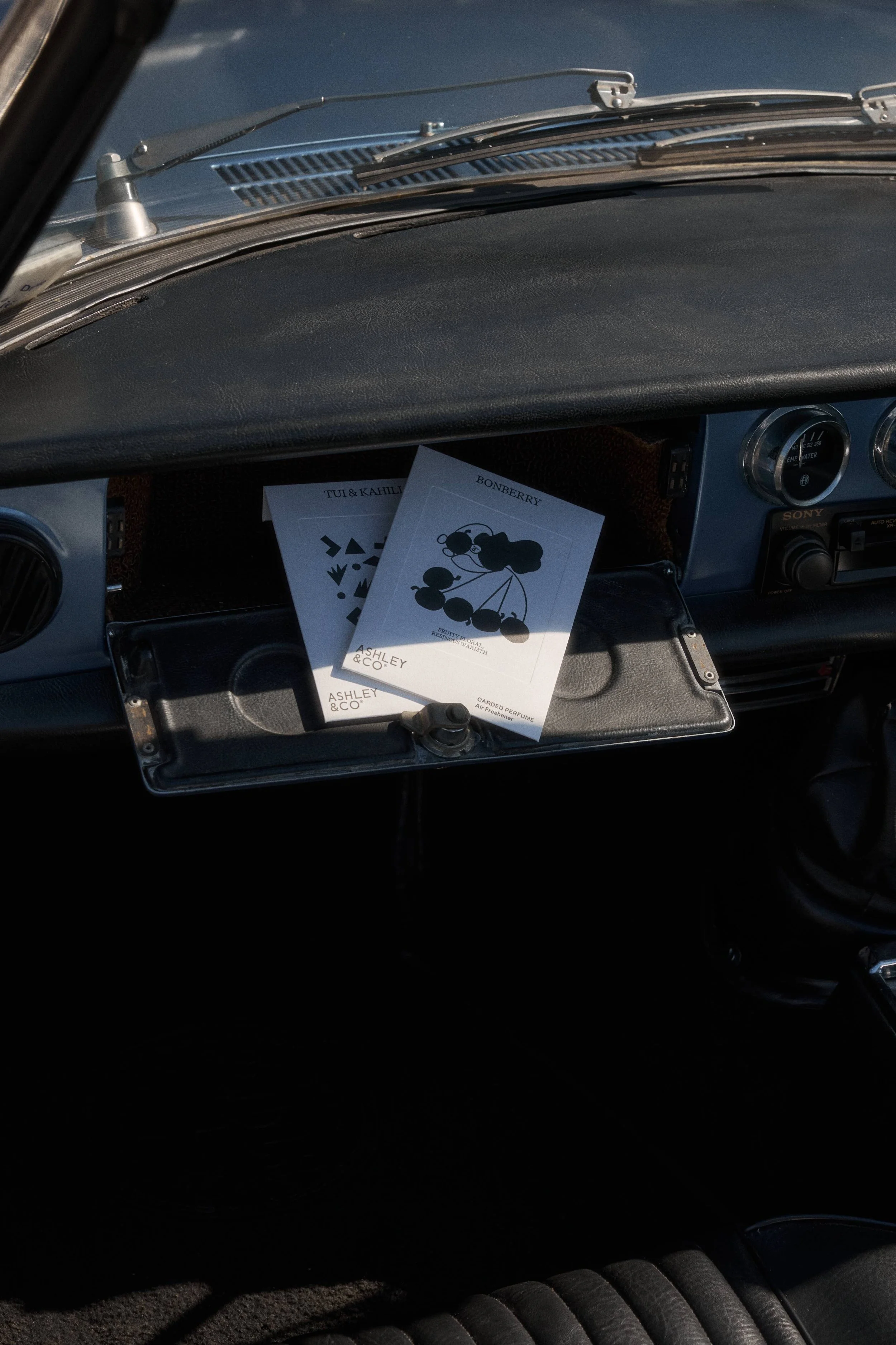 Inside a vintage car dashboard with two perfume sachets labeled 'Bonberry' and 'Tu & Kahlua' placed in a cubbyhole.