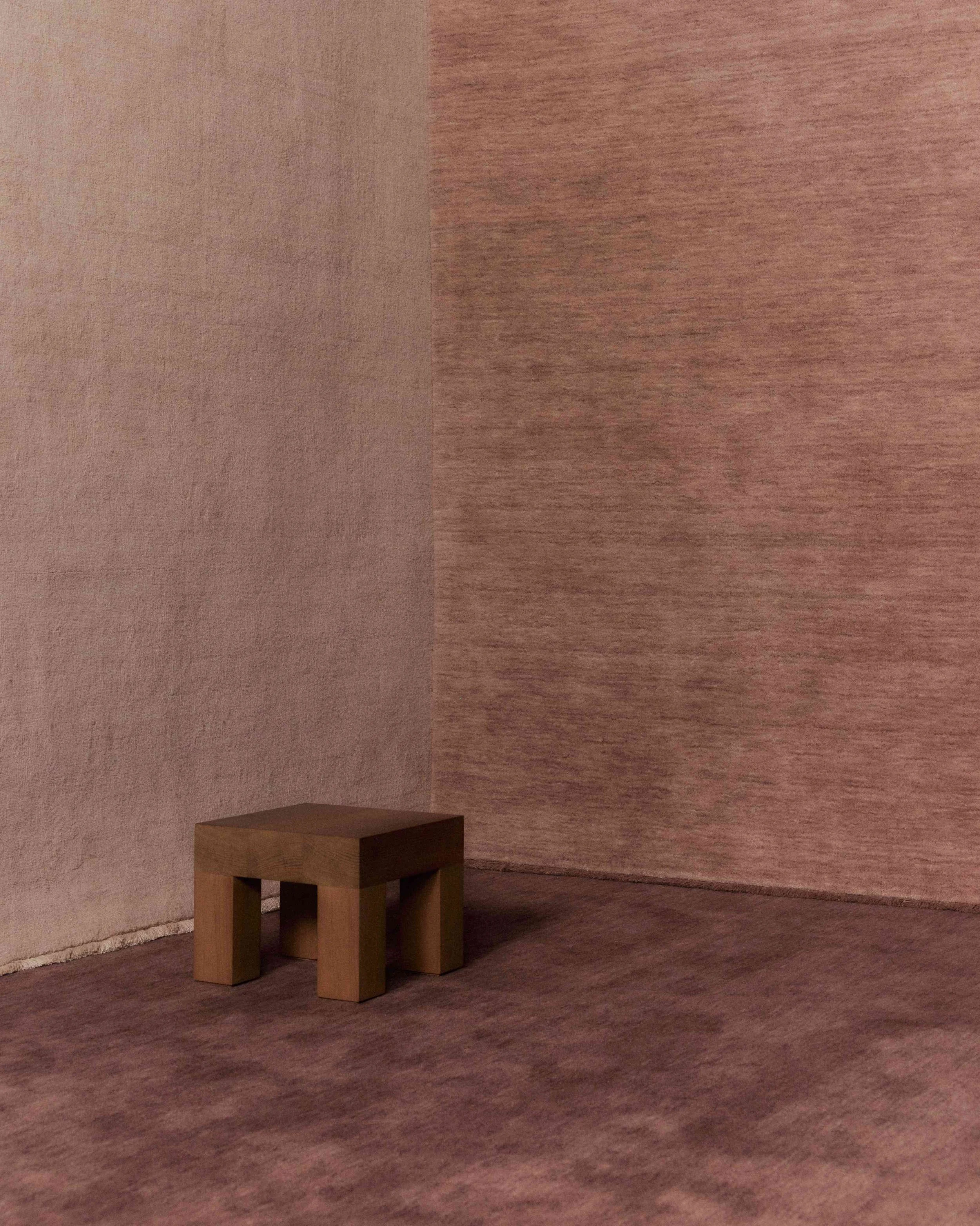 A small, wooden stool sits on a textured, pinkish-brown floor in a corner where two textured pink walls meet, made out of Baya rugs.