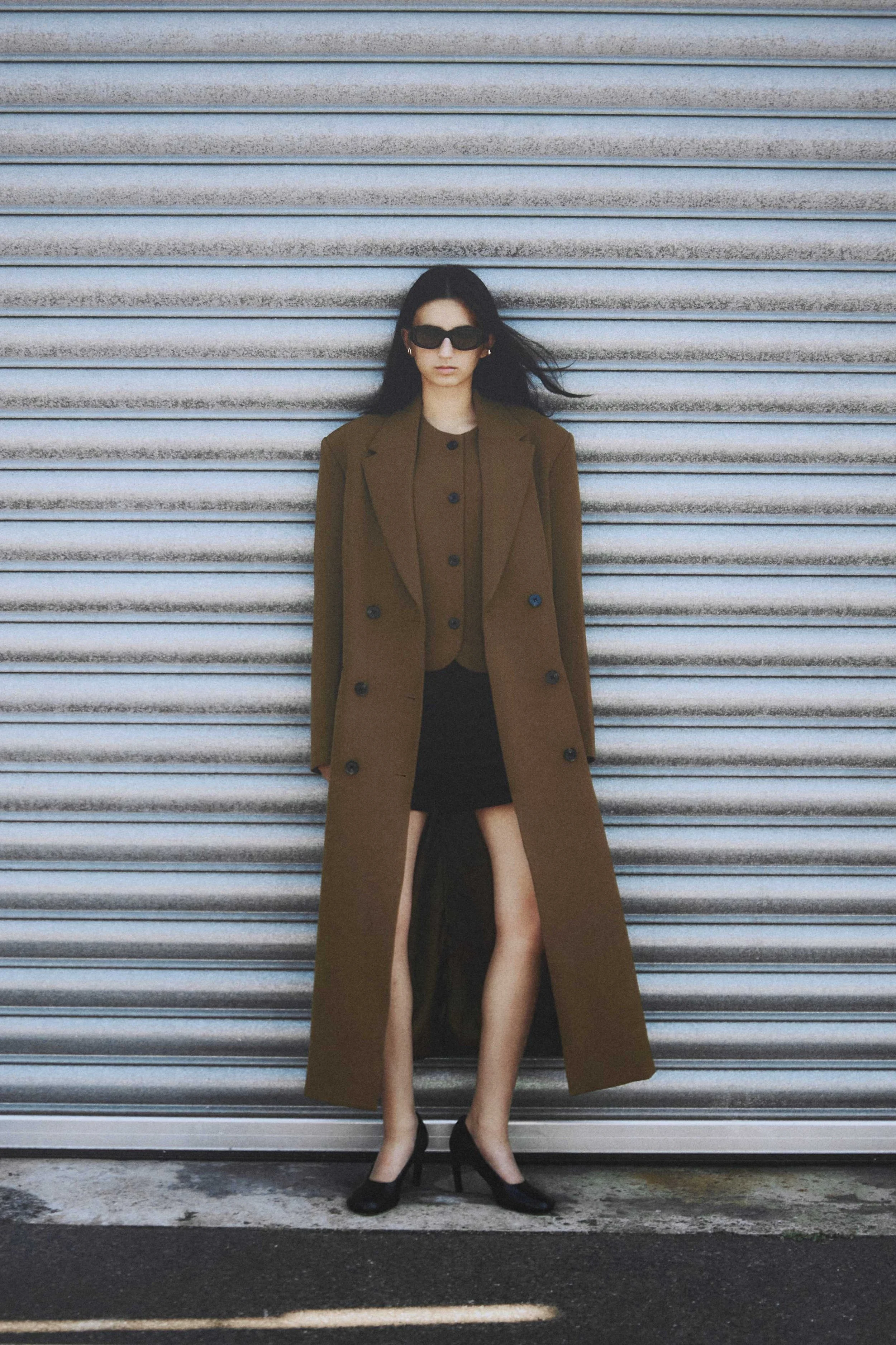 A woman with black hair wearing sunglasses, a brown long coat, a matching brown top, a black skirt with a high slit, and black high heels standing in front of a metal garage door.