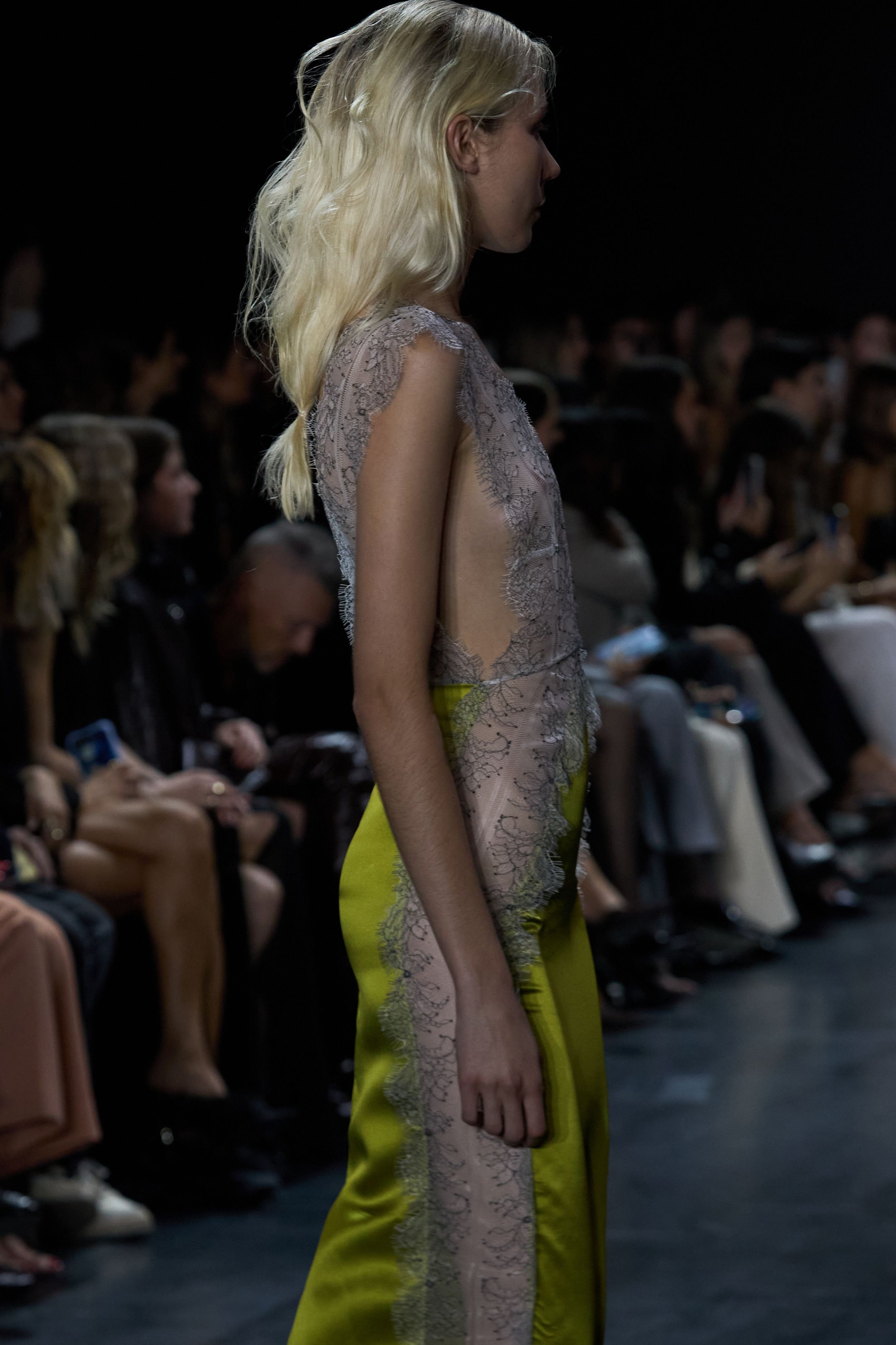 Fashion model walking down runway for Fashion Quarterly, in sheer lace top and bright yellow satin pants, with audience seated on either side.