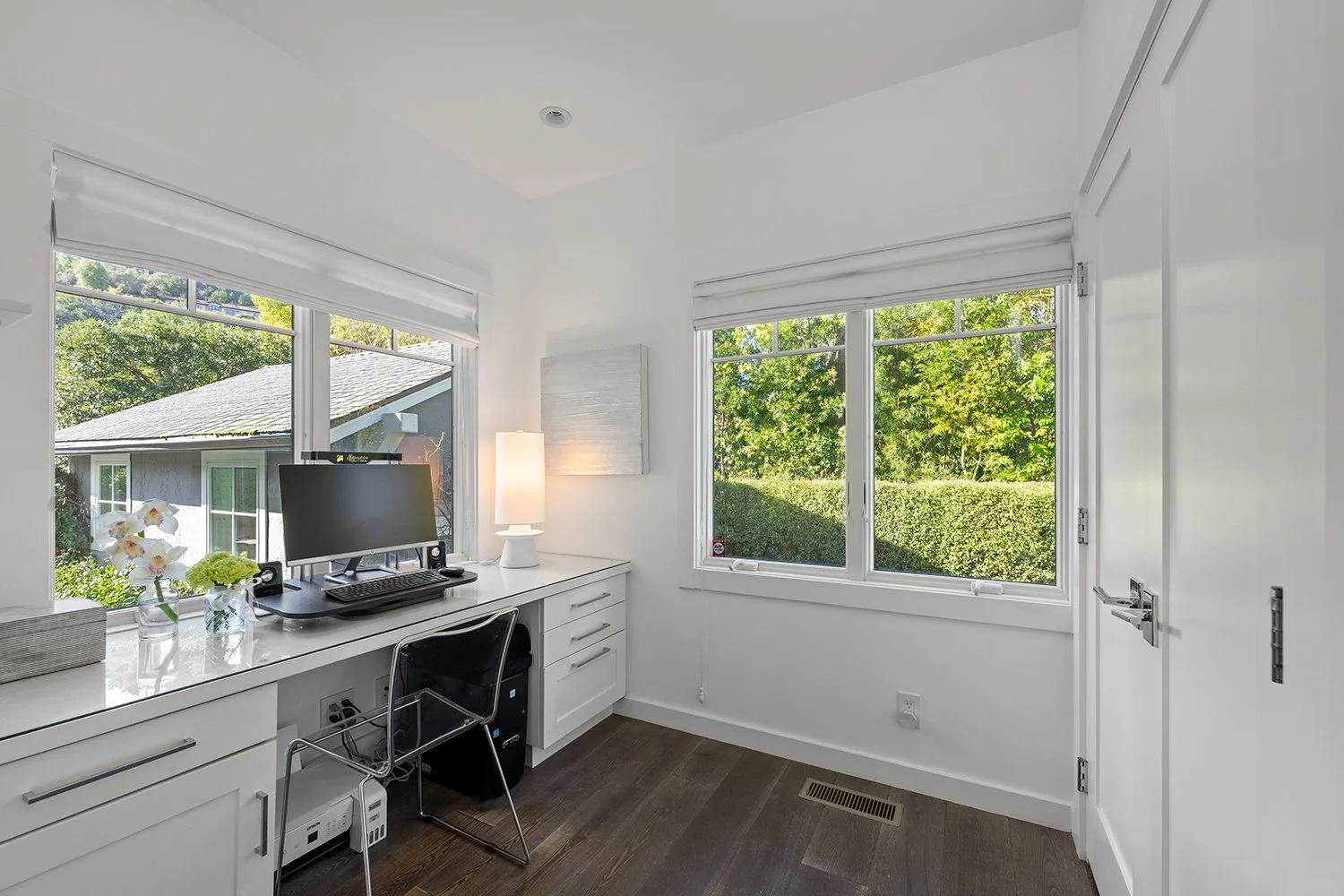 A bright home office with white walls, white cabinets, and dark hardwood flooring. A desk with a computer, flowers, and a lamp is located near large windows with white blinds, showing green trees outside.
