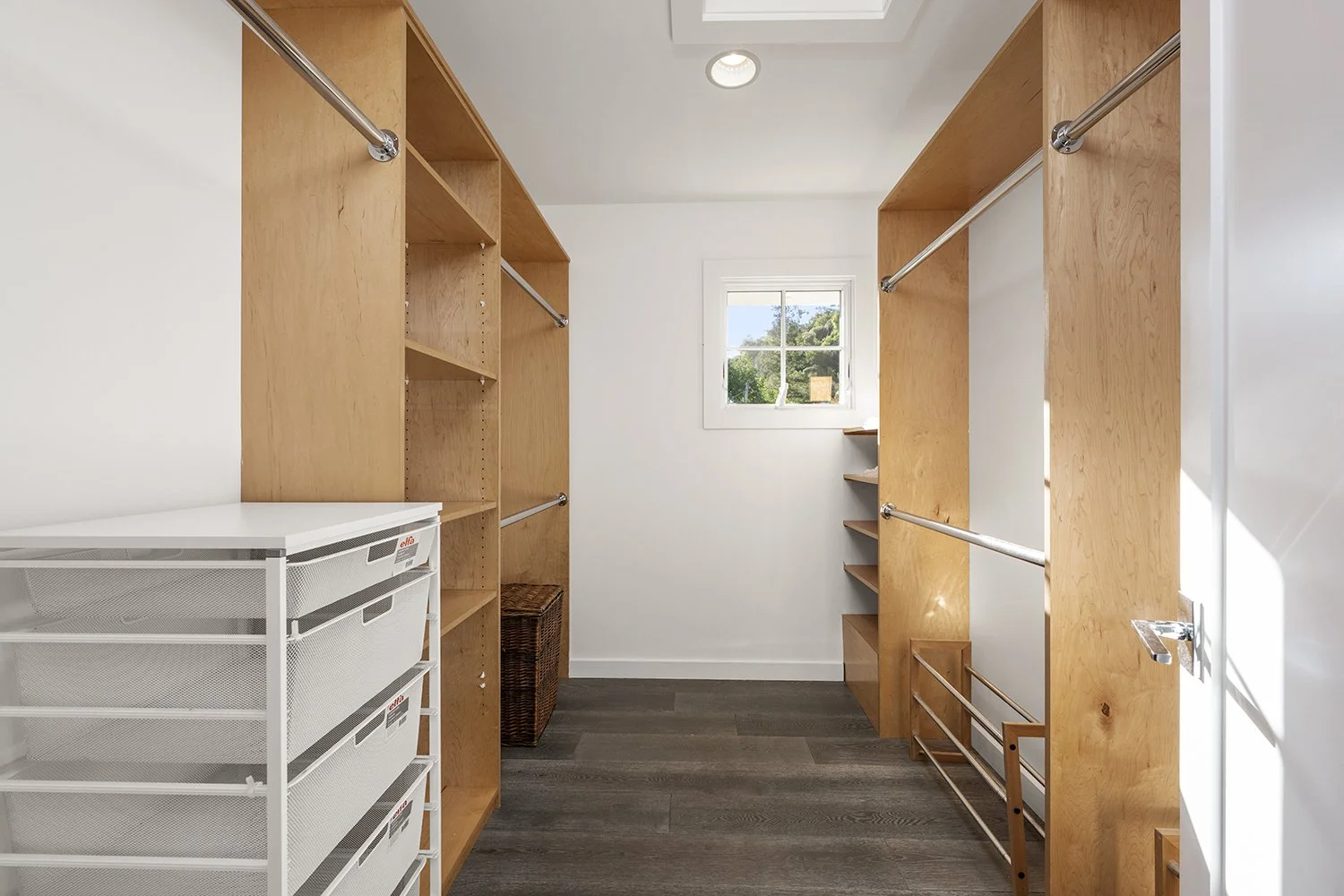 Empty walk-in closet with wooden shelves and rods, small window, white walls, and dark hardwood floor.