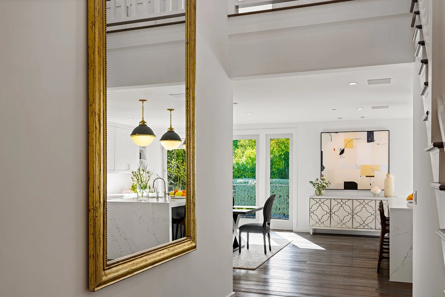 A bright, modern living space with white walls and natural light, featuring a gold-framed mirror, a white kitchen with marble countertops, two hanging pendant lights, a dining area with a dark table and chairs, and a white sideboard below a large abs