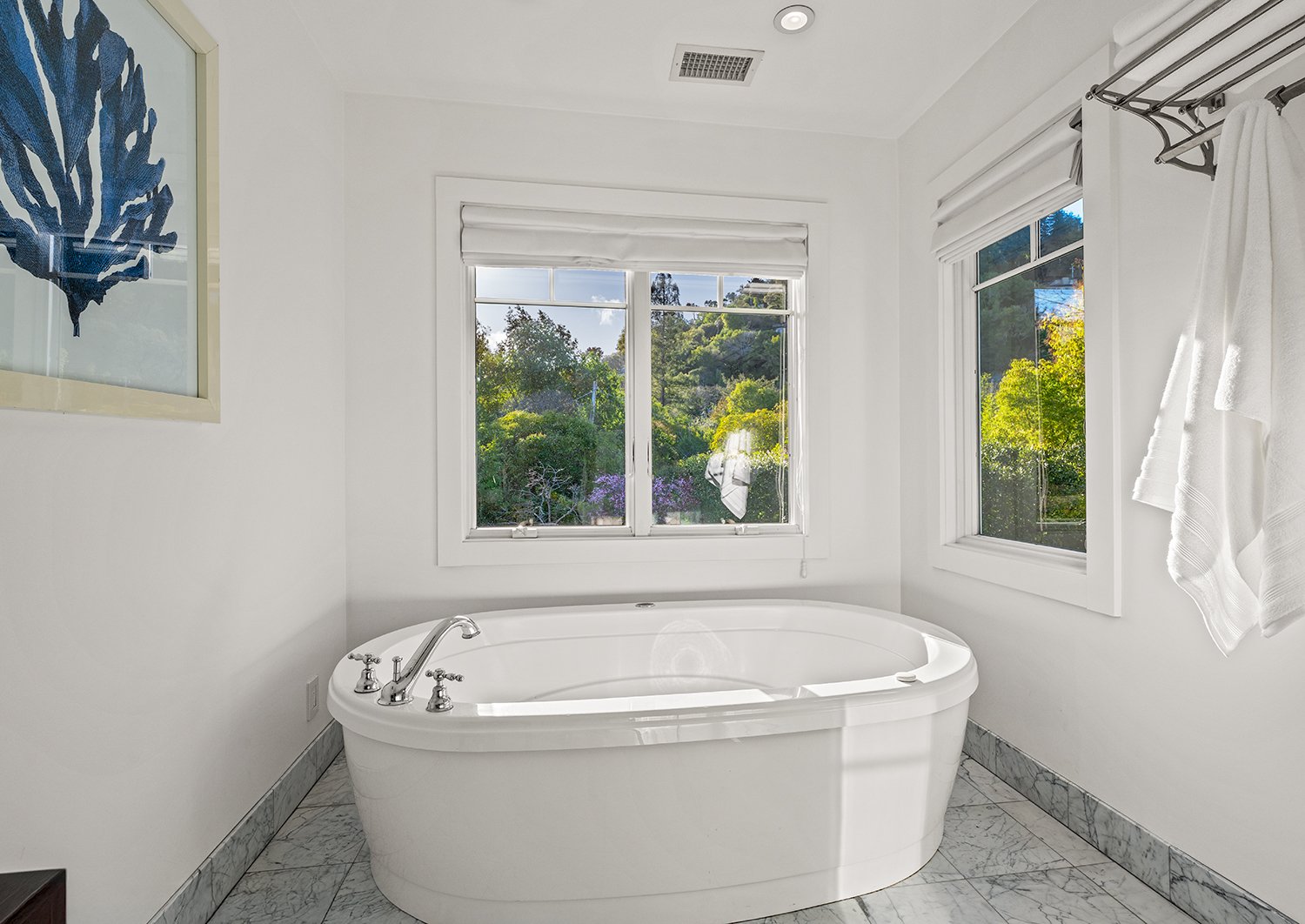 White bathtub in a bright bathroom with large windows and marble tile flooring.