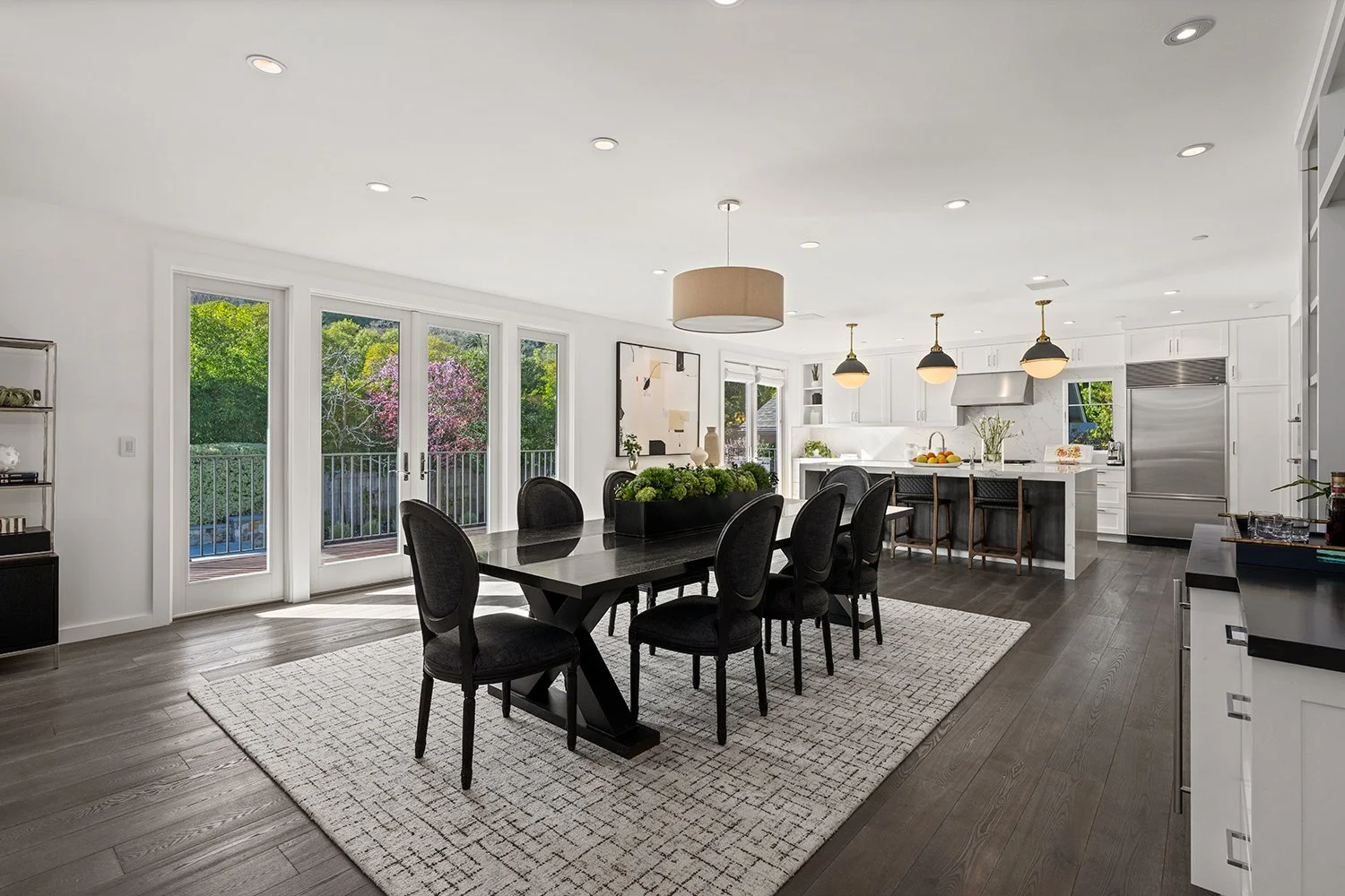 Open-concept kitchen and dining area with dark wood flooring, white cabinets, black dining table with six chairs, large windows, and sliding glass doors leading to a balcony with view of green trees.