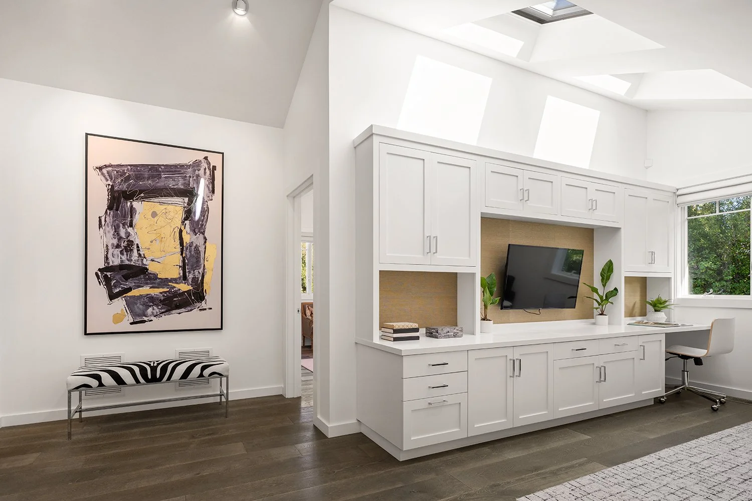 Modern living room with white cabinetry, a wall-mounted TV, plants, a patterned bench, and abstract artwork on the wall. Large windows and skylights provide natural light.