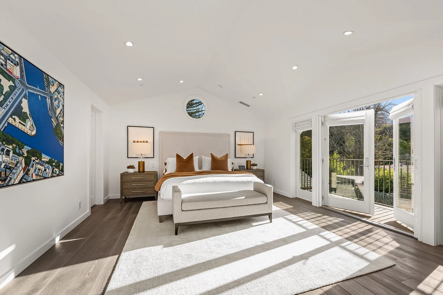 Bright bedroom with a large bed, nightstands with lamps, and large open glass doors leading to a balcony with a view of greenery.