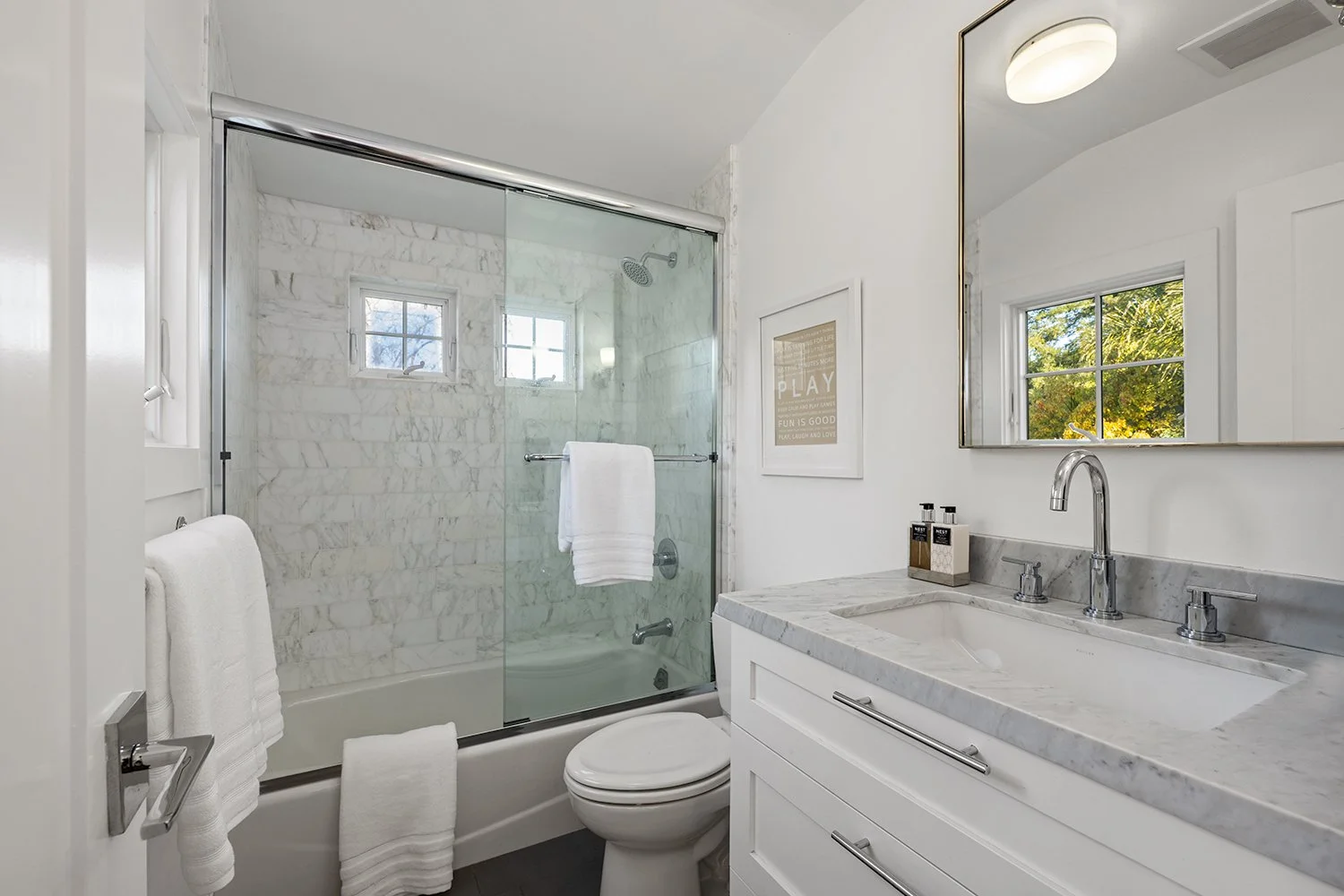 A modern bathroom with a shower area featuring white marble tiles, two small windows, a glass sliding door, a white toilet, a white vanity with a marble countertop, a large mirror, and a window showing outdoor trees.