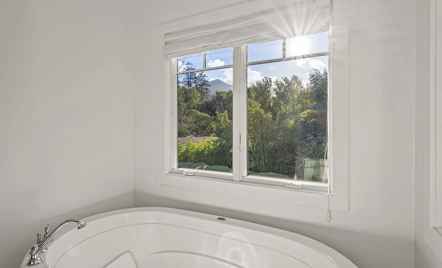 Bathroom with a large window overlooking a lush green landscape with trees and mountains in the distance, sunlight shining through the window.