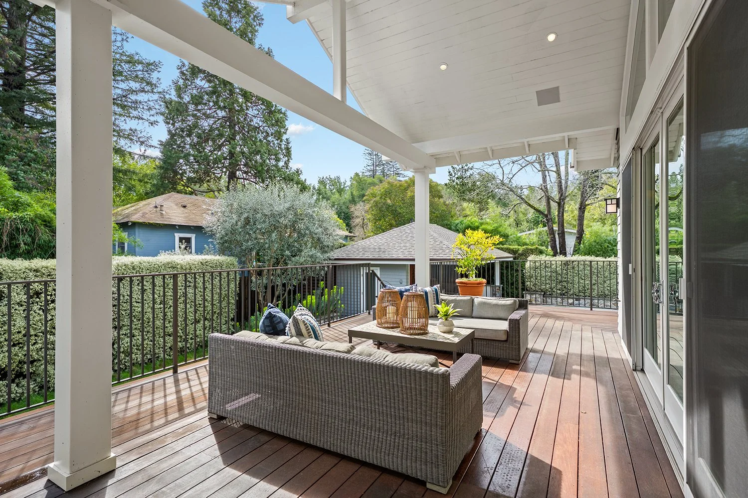 An outdoor patio with wicker furniture, including a sofa and chairs, decorated with pillows. There is a wooden table with lanterns and a potted plant. The patio has a wooden floor, black railing, and is shaded by a white ceiling. Trees and neighborin