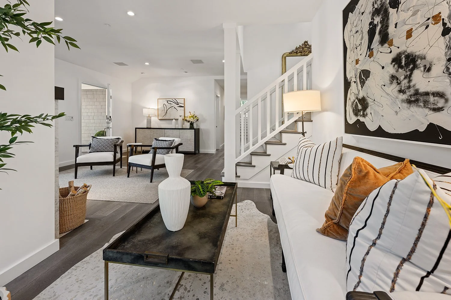 Living room with white walls and modern decor, featuring a white sofa with patterned pillows, a black coffee table with a vase and plants, a staircase with white railing, and art on the walls.
